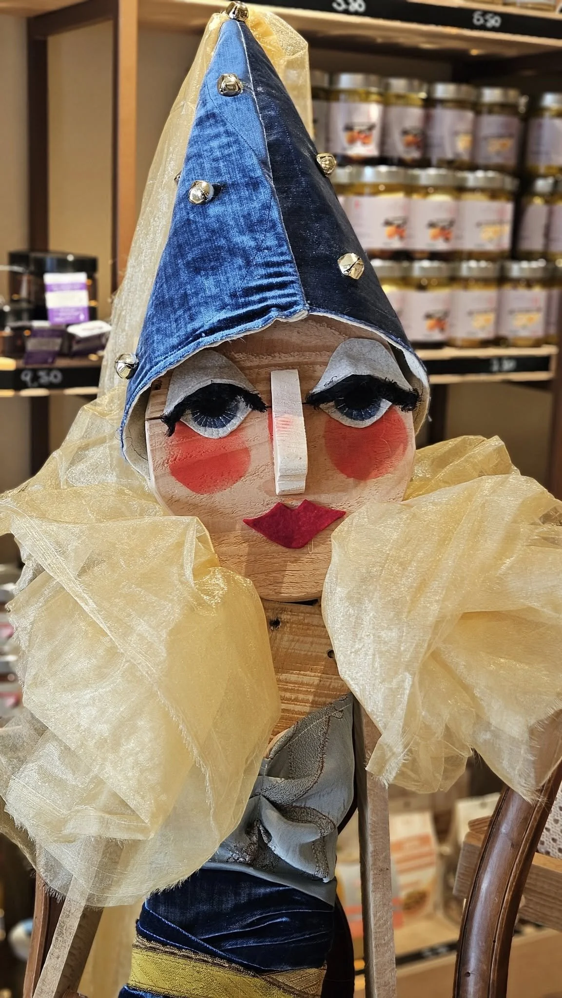 Statua o marionetta con volto stilizzato, dettagli per occhi, bocca, e naso, tessuto blu sulla testa, tessuto giallo chiaro intorno al collo, e parti in tessuto beige e altri materiali, posizionata in negozio.