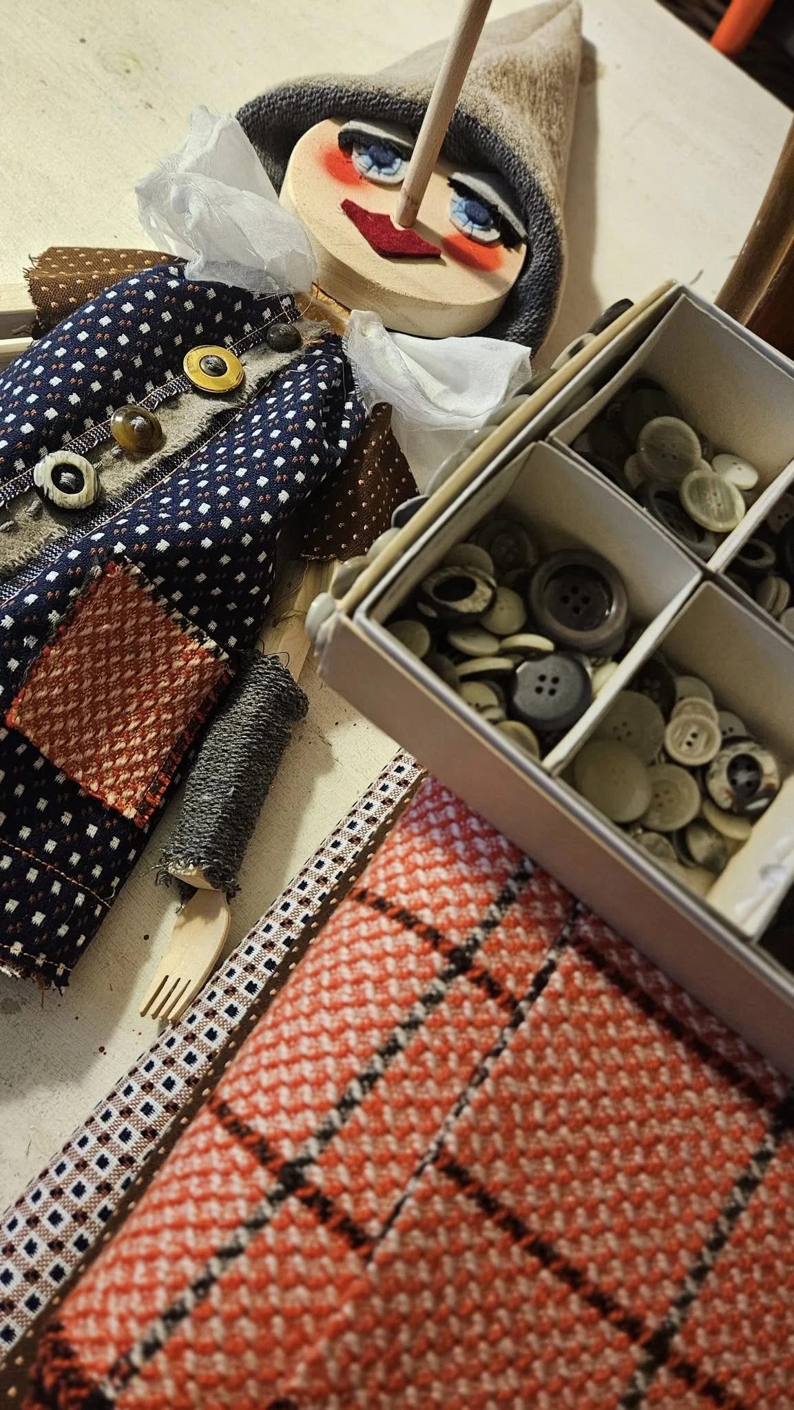 Un manichino con abbigliamento colorato, un volto di stoffa con accanto un box con bottoni di vari colori e forme, e una coppia di braccia e mani di stoffa con dettagli decorativi.