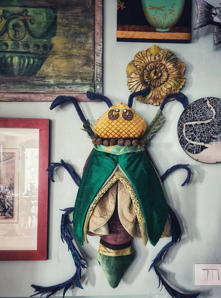 Decorazione murale artista in forma di insetto con corpo verde, testa gialla con occhi e bocca, zampette blu, e dettagli floreali e ceramici sul muro.