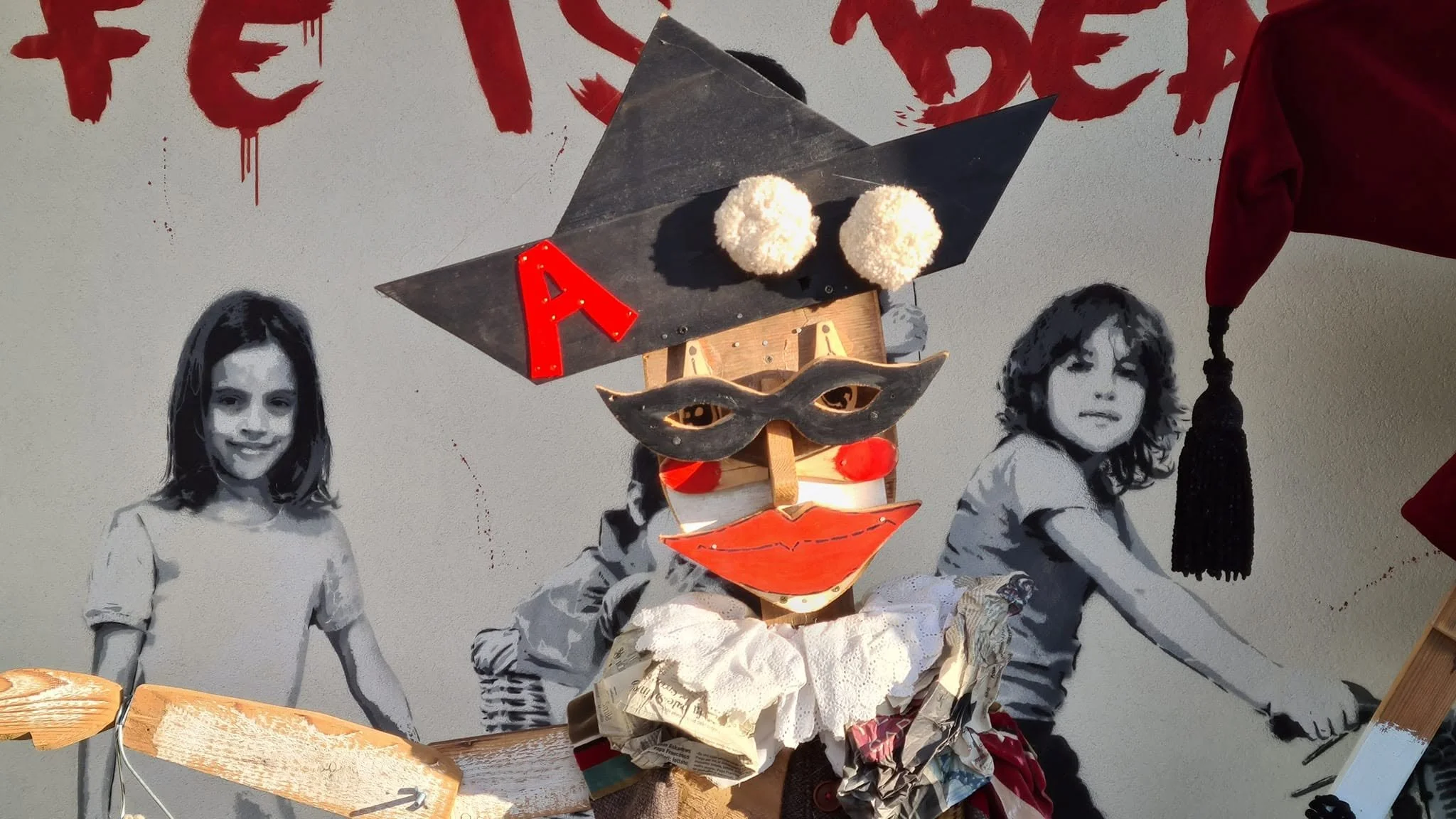 Un'immagine di un costume di Carnevale fatto a mano con una maschera, due palline di peluche sulla testa, e decorazioni di carta e stoffa, circondato da figure di bambini disegnate in bianco e nero.
