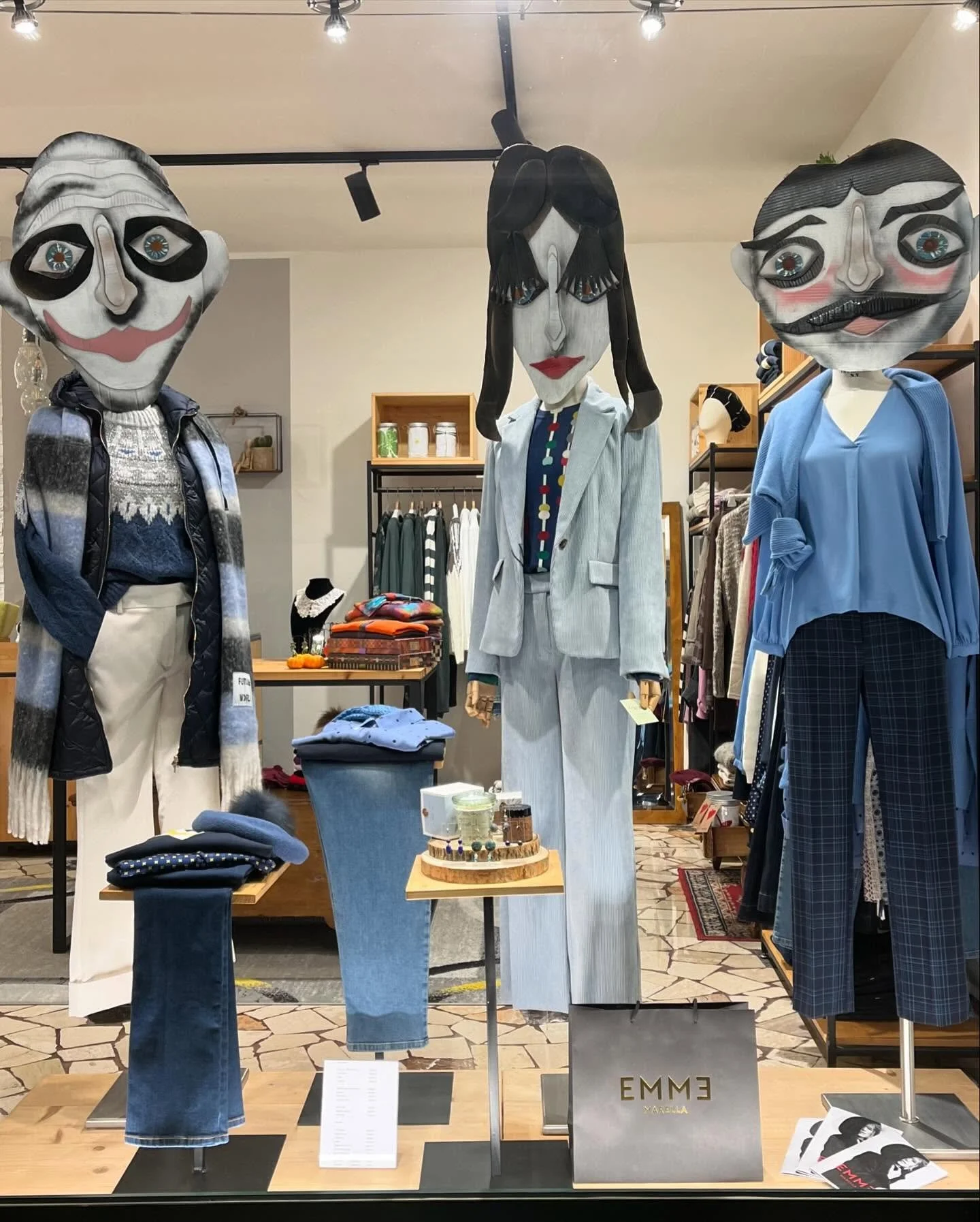 Vetrina di negozio di abbigliamento con tre manichini con teste di cartapesta con volti artistici; uno con capelli neri e un vestito a righe, uno con una giacca blu ed un pantalone a scacchi, e uno con una giacca a quadri. In primo piano ci sono magl