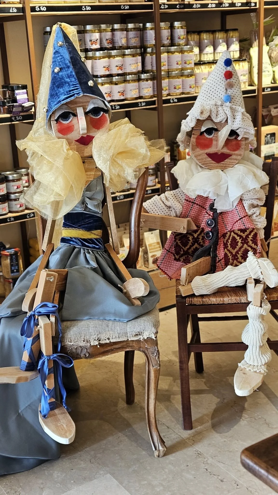 Due figure di marionette in legno vestite con abiti colorati e cappelli decorati, sedute su sedie di legno in un negozio. Sullo sfondo scaffali con barattoli e prodotti alimentari.