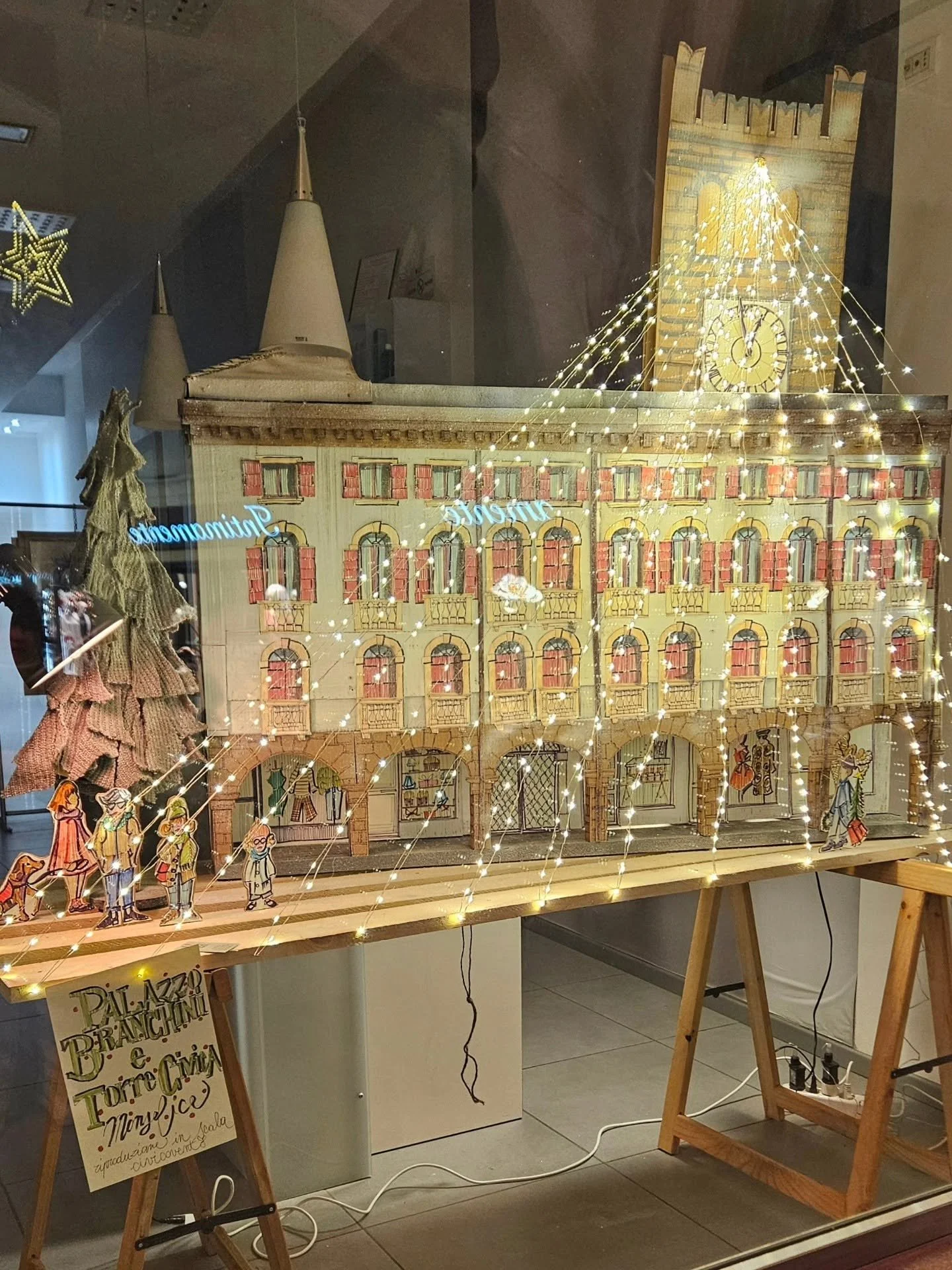 Decorazione natalizia con una ricostruzione di un edificio e un castello, luci luminose, figure di cartone animate di persone, e un cartello che pubblicizza pizza, panini, e altro.
