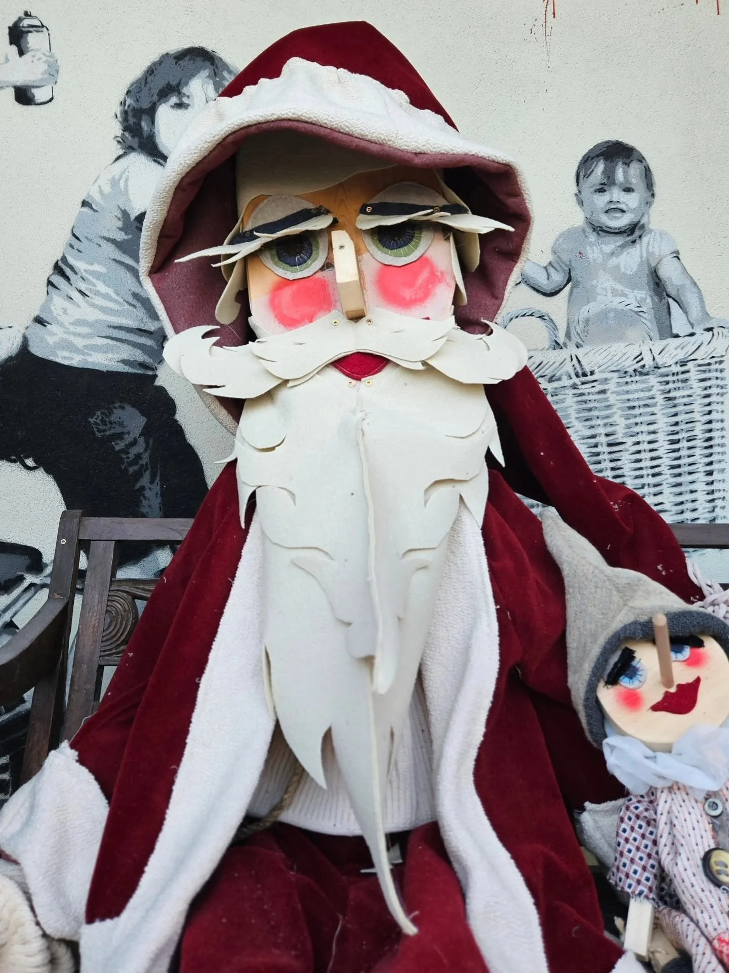 Babbo Natale con barba e occhi grandi, vestito di rosso e cappello, seduto davanti a un murale con bambini.