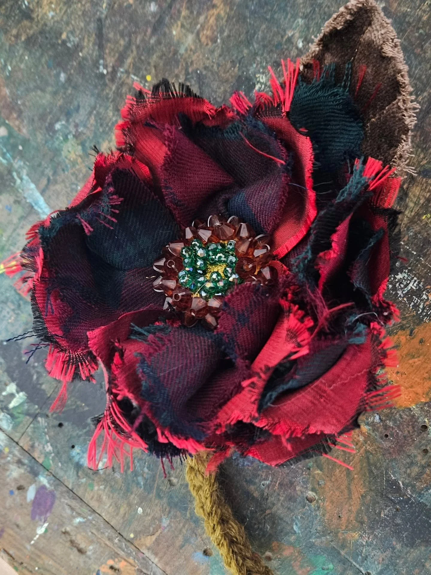 Fiore artigianale con bottoni di perline decorative, avvolto da tessuto scozzese rosso e nero, attaccato a un cordino di spago di colore marrone. Sullo sfondo, superficie di legno decorata con macchie e segni di vernice.