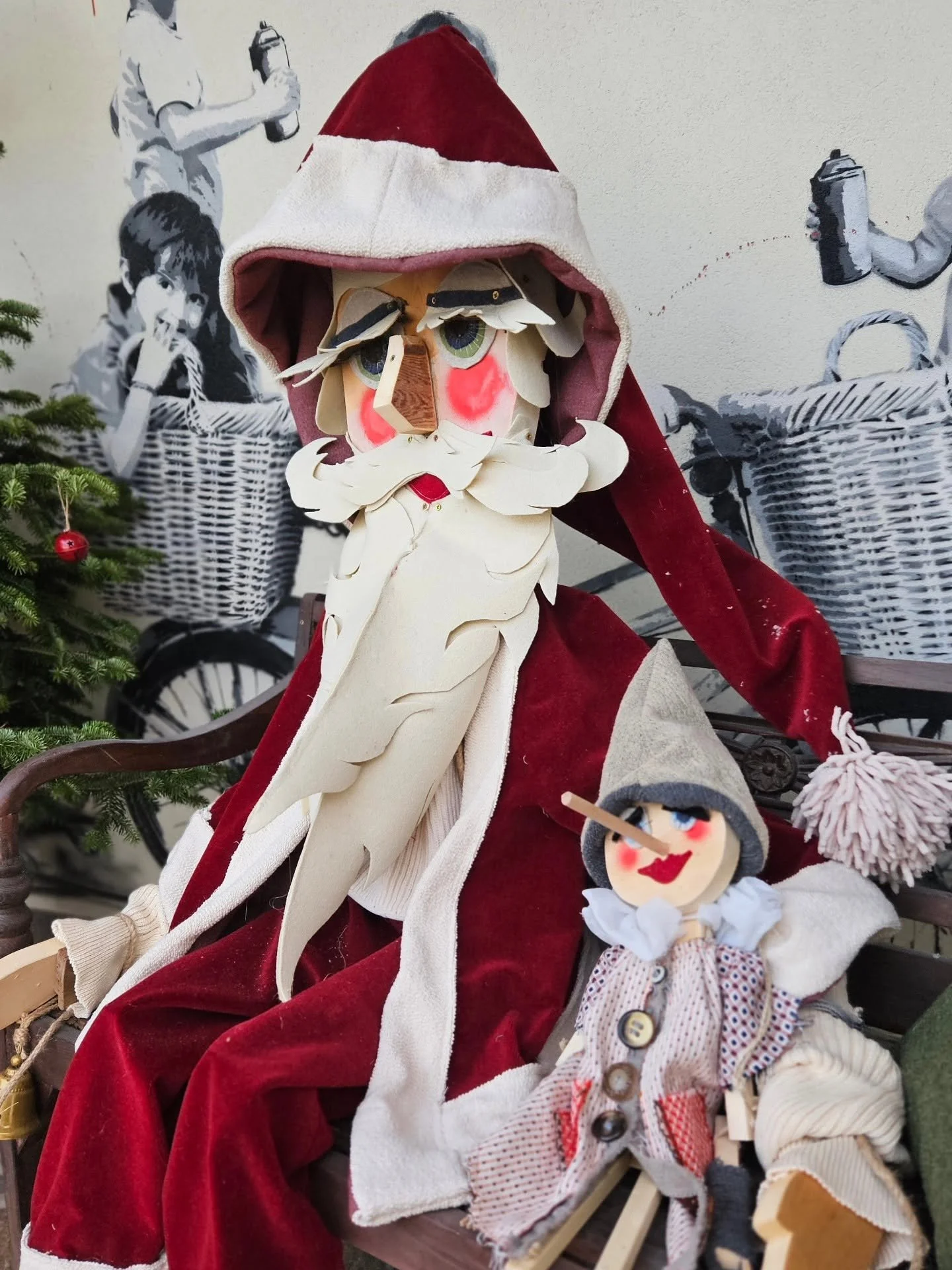 Pupazzo di Babbo Natale e bambola, entrambi con abiti natalizi, seduti su una panchina davanti a un muro con decorazioni natalizie.