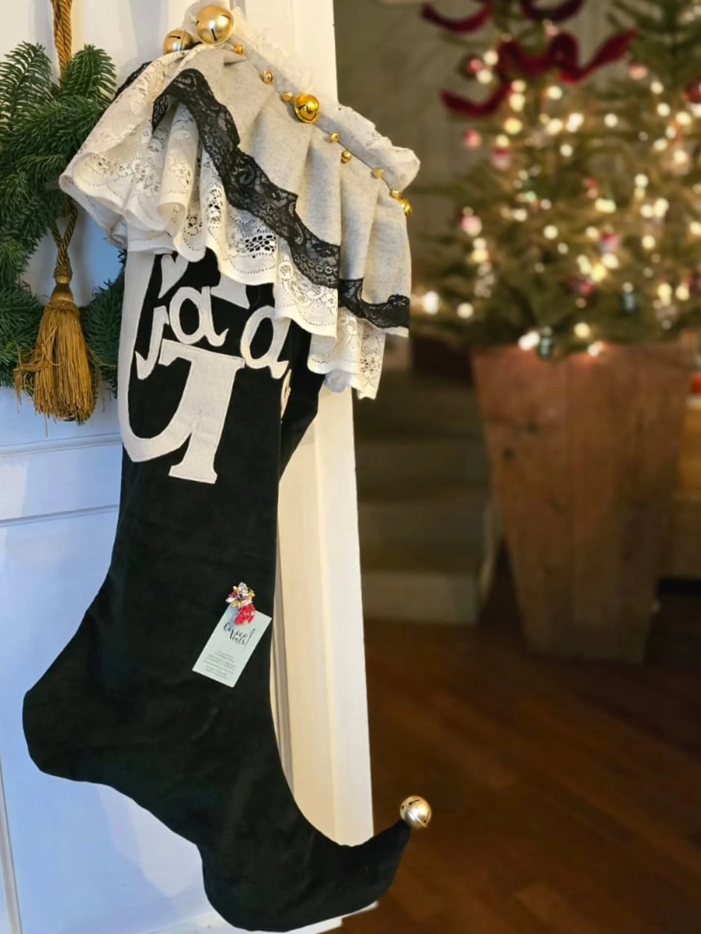 Decorazione natalizia con calza natalizia decorata con pizzi bianchi e neri, perni dorati e palline dorate, attaccata a un angolo di una parete, con un albero di Natale sullo sfondo decorato con luci e palline colorate.