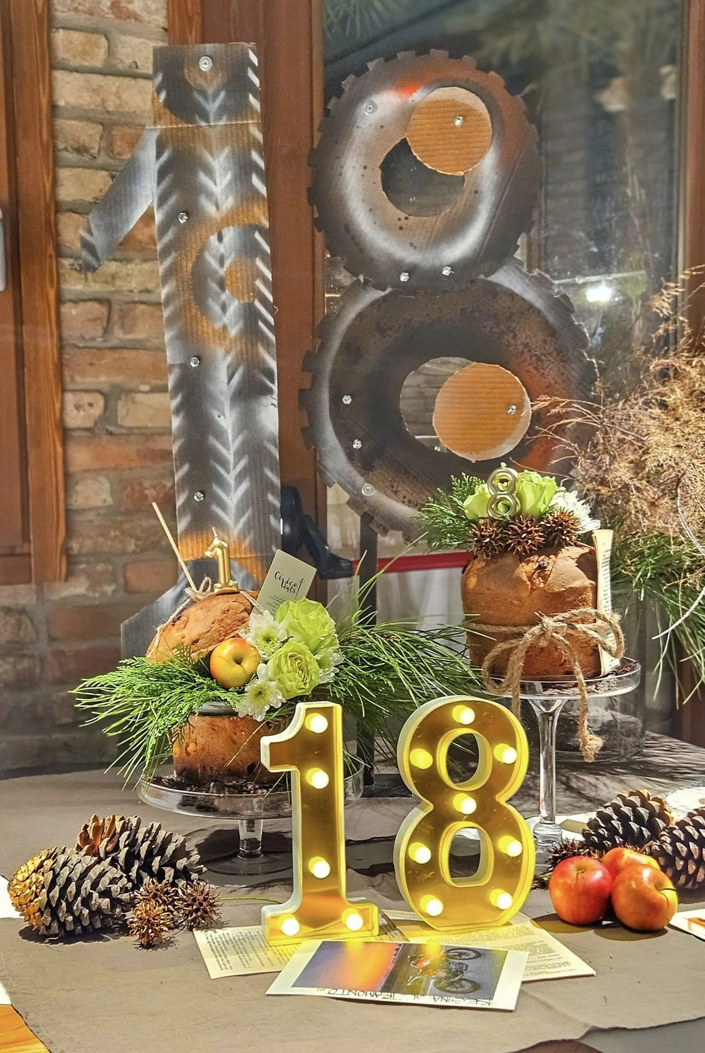 Decorazione di festa con cioccolata, mele e rami di pino, numeri luminosi 1 e 8, sfondo con motore di bicicletta e pareti di mattoni.