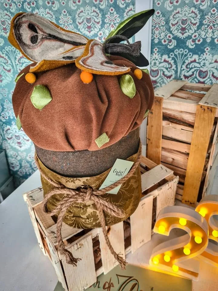 Collana di cappelli di stoffa decorati con foglie e fiori, esposta su una confezione di legno con sfondo di carta da parati colorata.