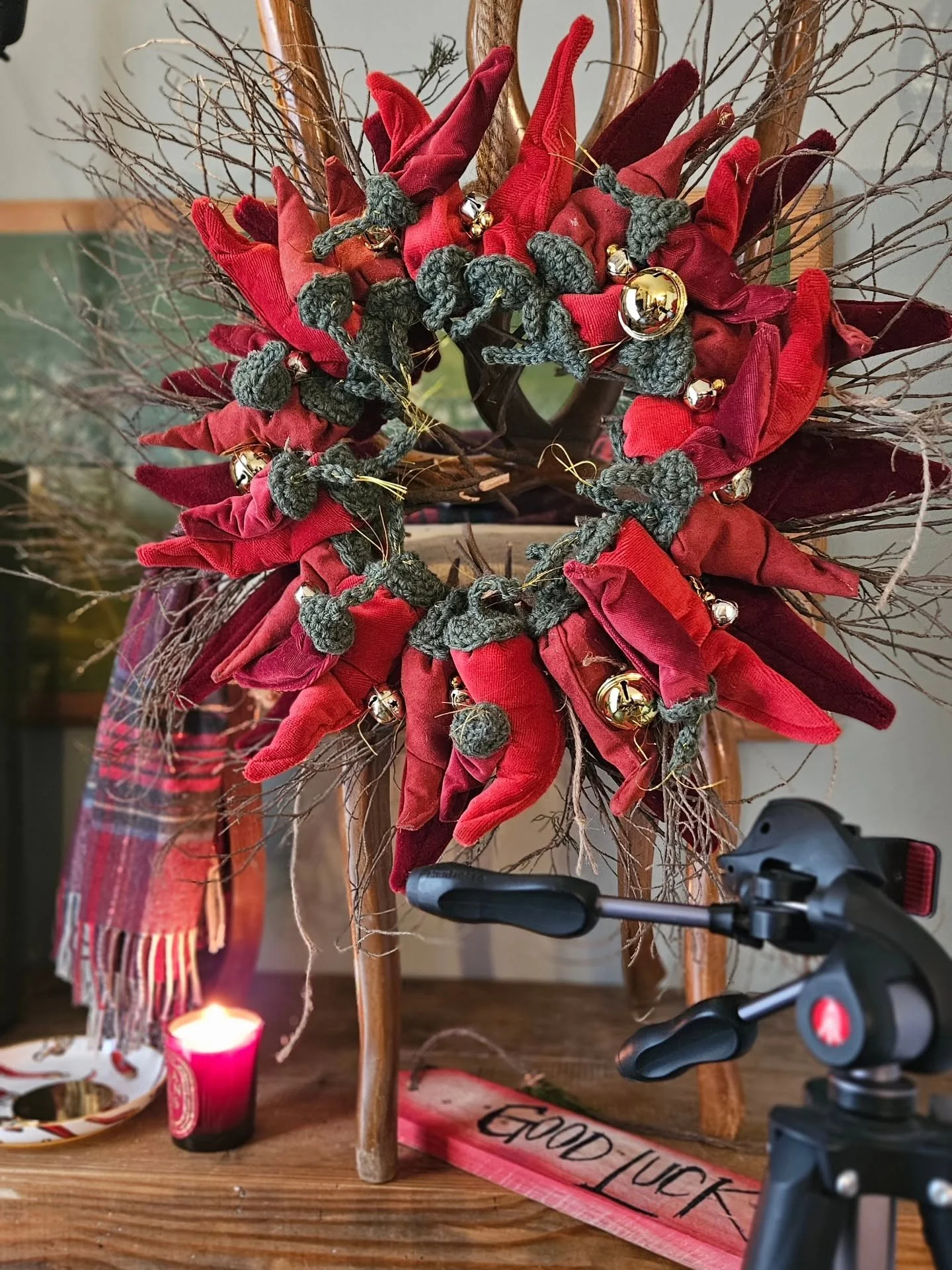 Decorazione natalizia con corone di stoffa rosse, rametti secchi e pigne, lampada a candela rosa e bastone decorativo con scritta 'Good Luck' su un tavolo di legno.