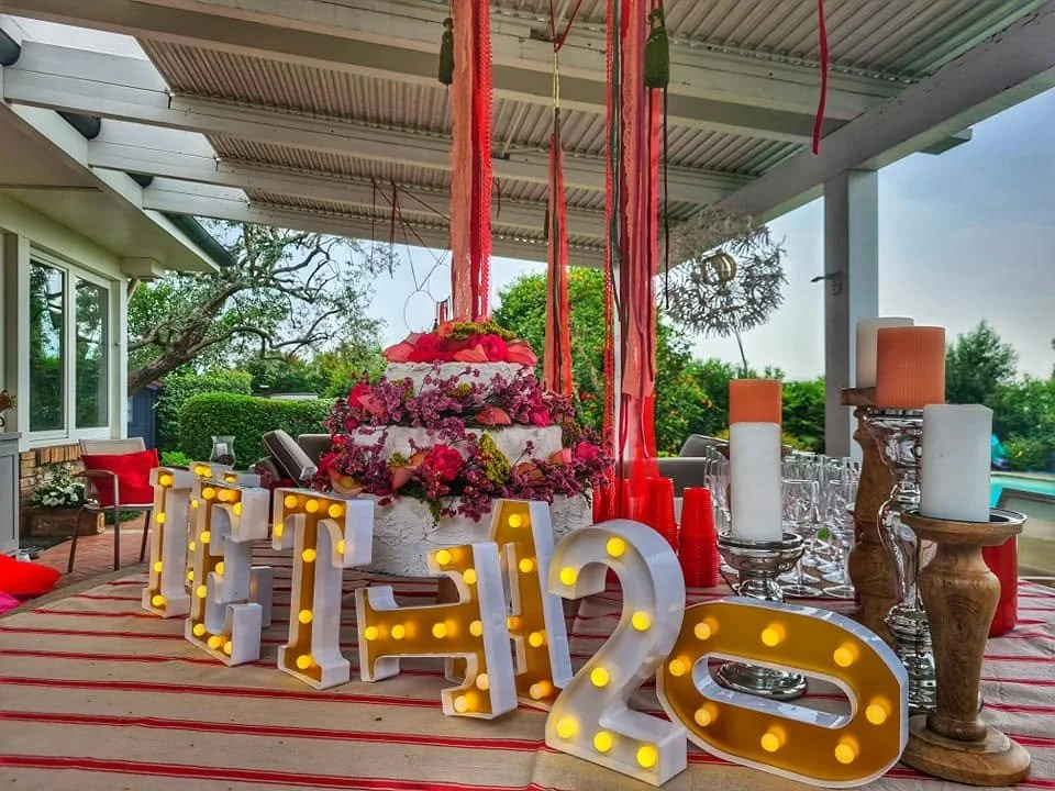 Decorazioni per una festa di compleanno con la scritta luminosa 'TANTI 2O' davanti a un grande torta decorata con fiori e frutta, su un tavolo coperto con un tovagliato rosso e bianco, con candele e bicchieri trasparenti.