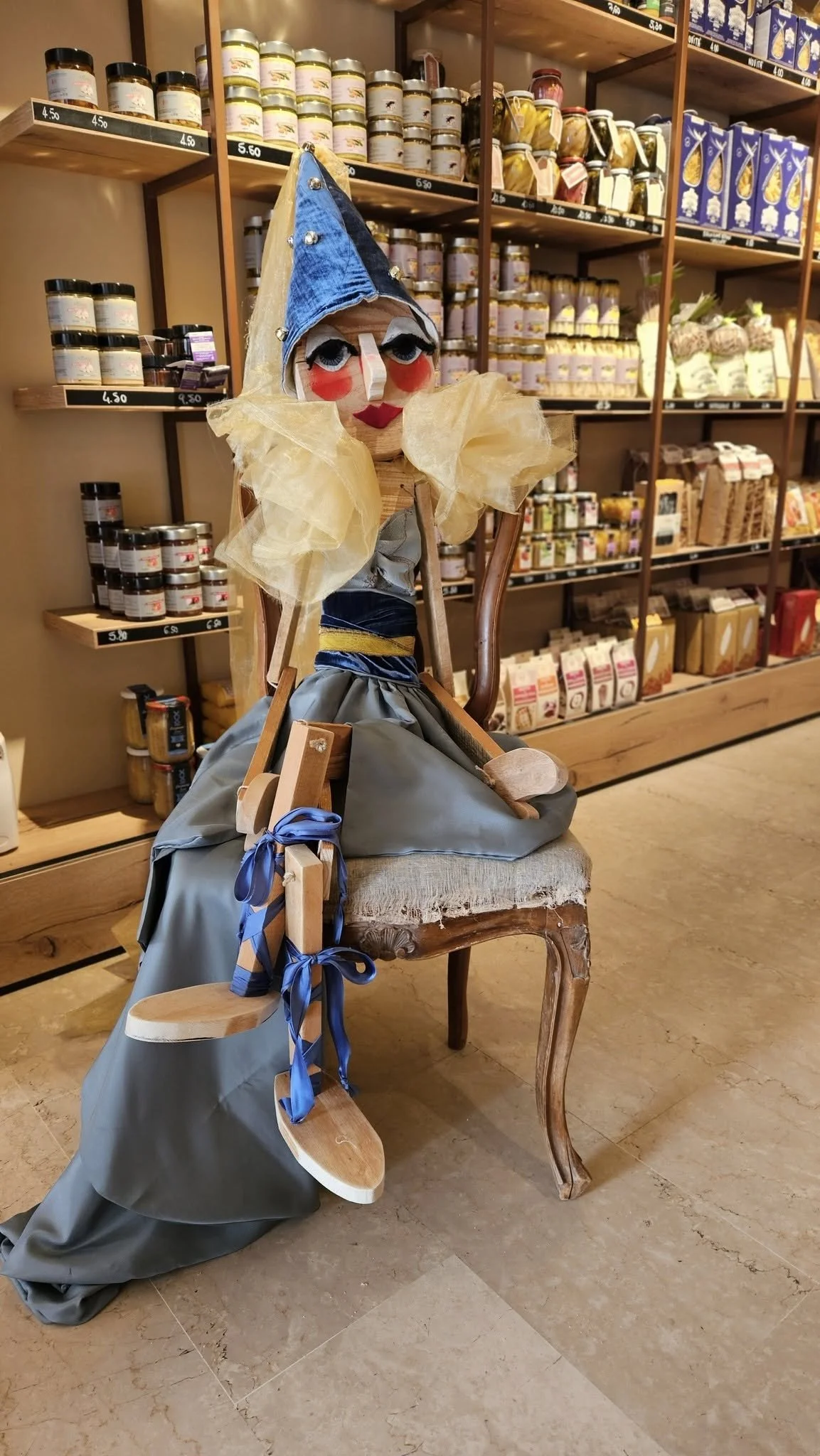 Una bambola di legno con abiti di stoffa, seduta su una sedia di legno rustica, in un negozio di alimentari con scaffali pieni di conserve, biscotti e pasta.