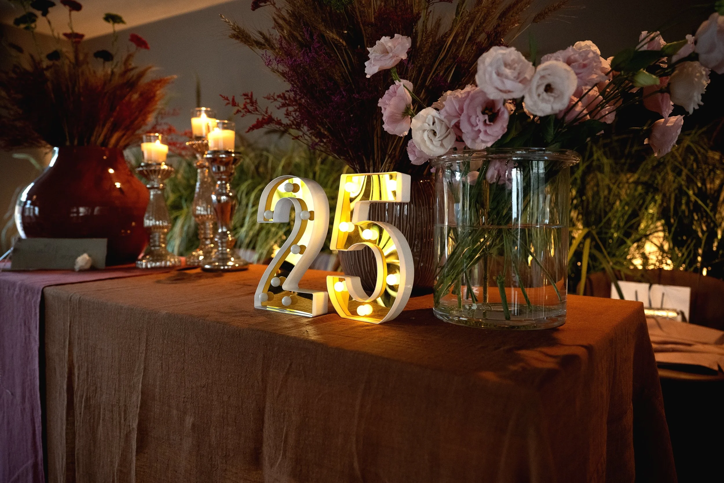 Decorazione di una tavola con vasi di fiori, candele accese e un'illuminazione calda, con un numero 25 illuminato come decorazione. Ambiente elegante e intimo.
