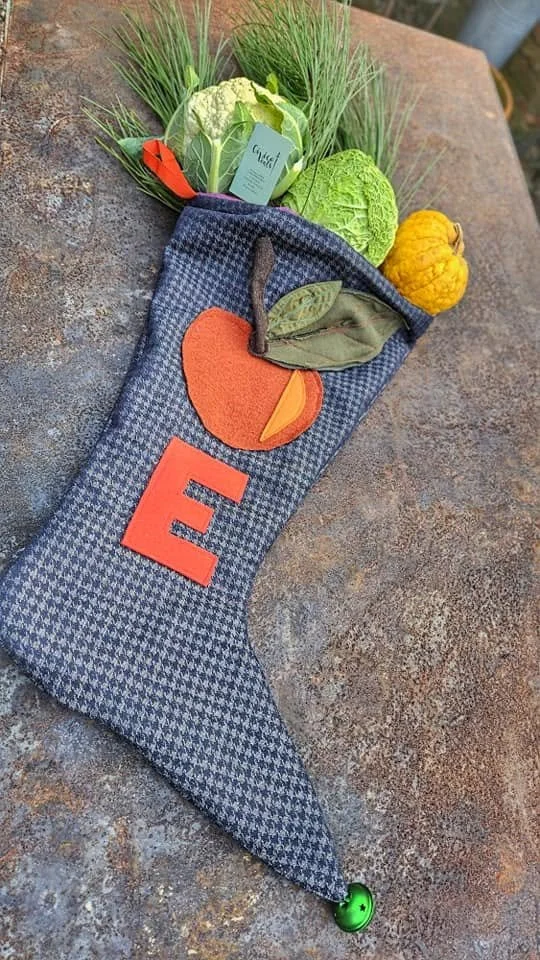 Calza natalizia decorata con frutta di feltro, contenente vari ortaggi e verdure realizzate con materiali tessili, posizionata su una superficie di pietra.