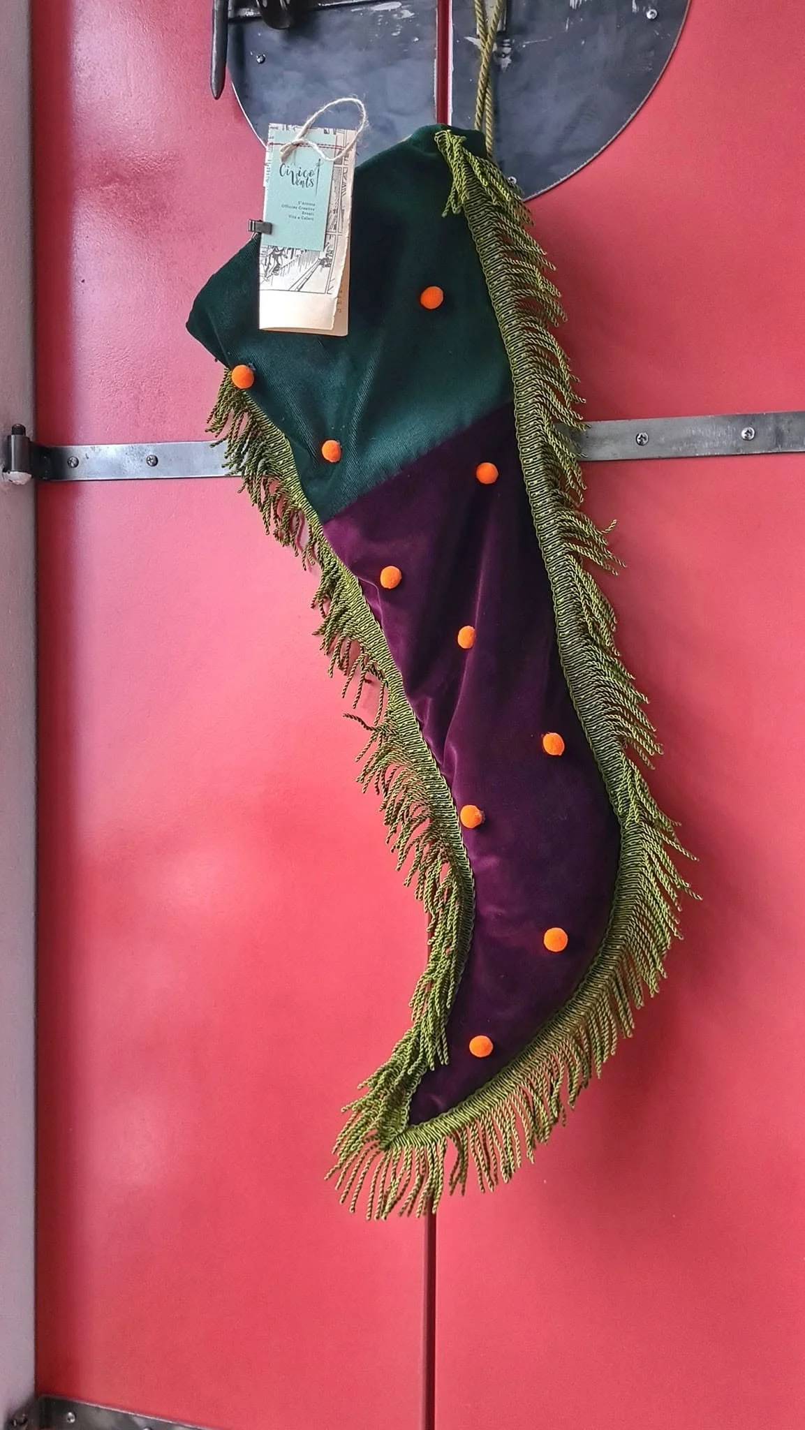 Una calza della Befana colorata, decorata con pompon arancioni e frange verdi, appesa su una porta rosa di Civico vent8