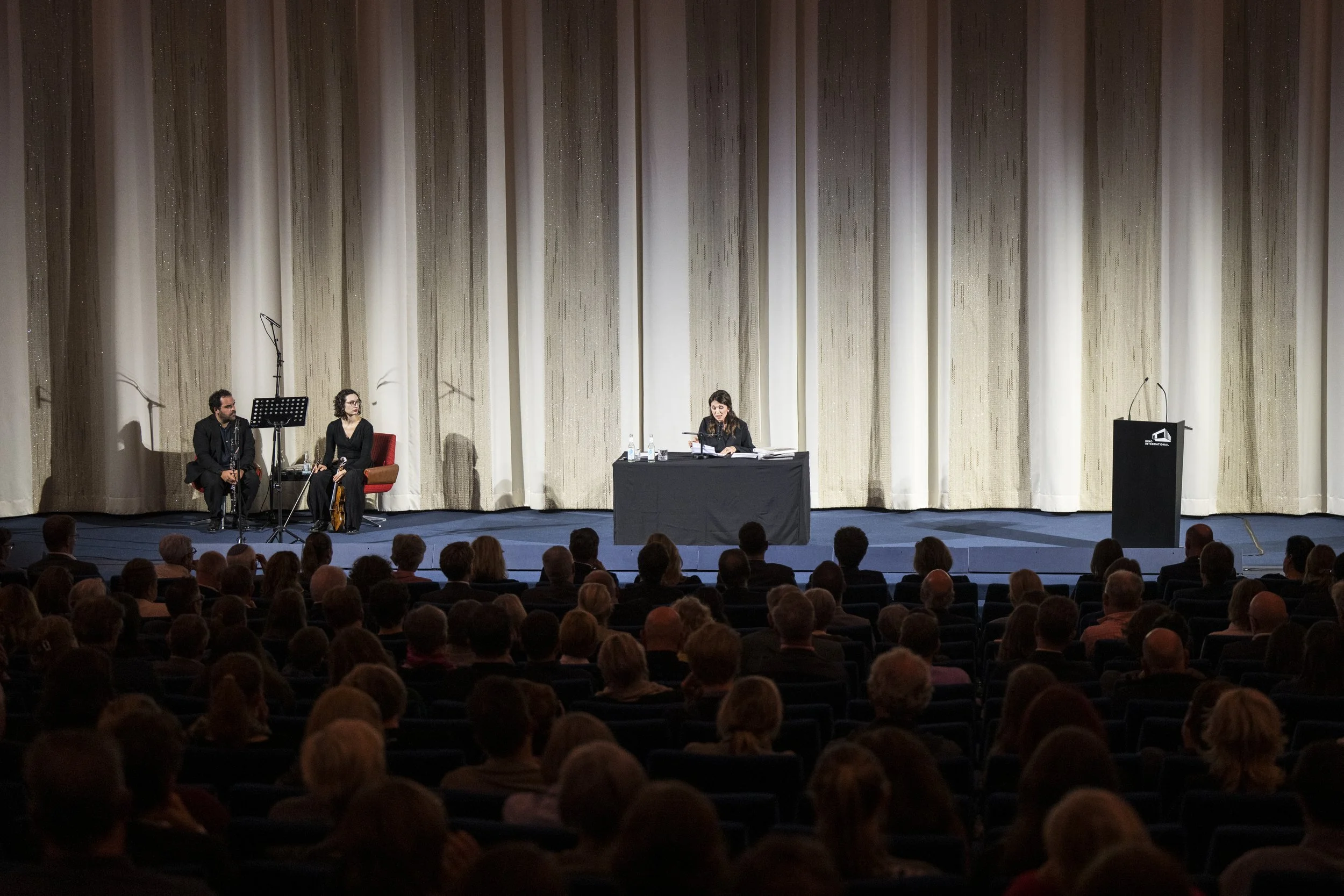 A panel discussion on stage with three women, two men, microphones, water bottles, and a podium, in front of a large curtain, audience seated in the theater.