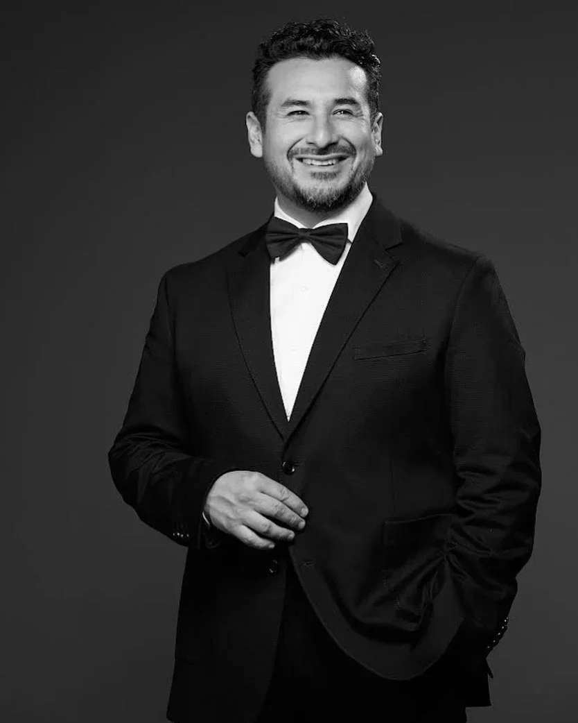 Black and white portrait of a man in a tuxedo with a bow tie, smiling, with one hand adjusting his jacket, against a dark background.