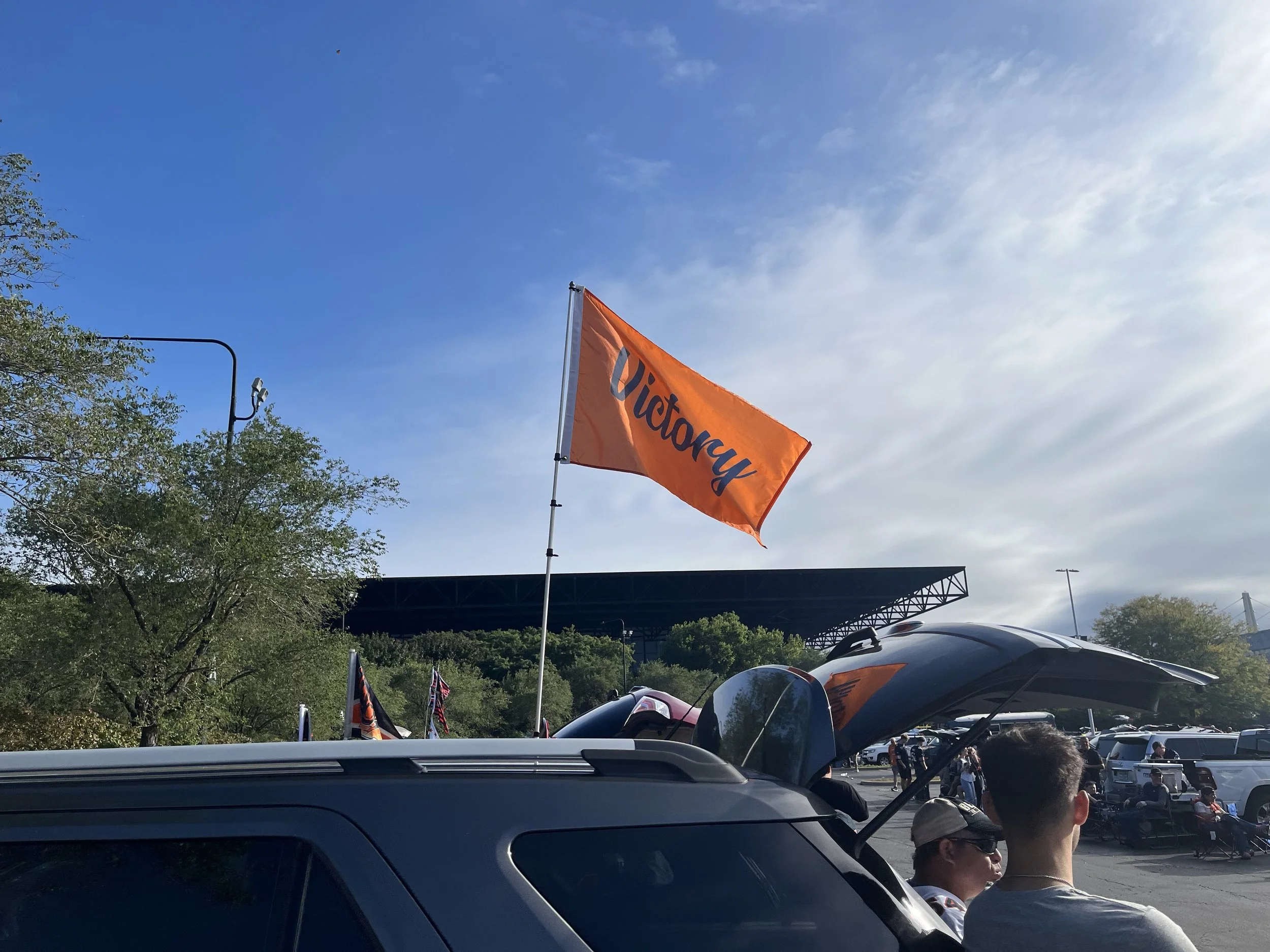 People gather at an outdoor parking lot with cars, some with open trunks. An orange flag with the word "Victory" flies on a pole. The sky is partly cloudy with some blue visible, and there are trees and a stage in the background.