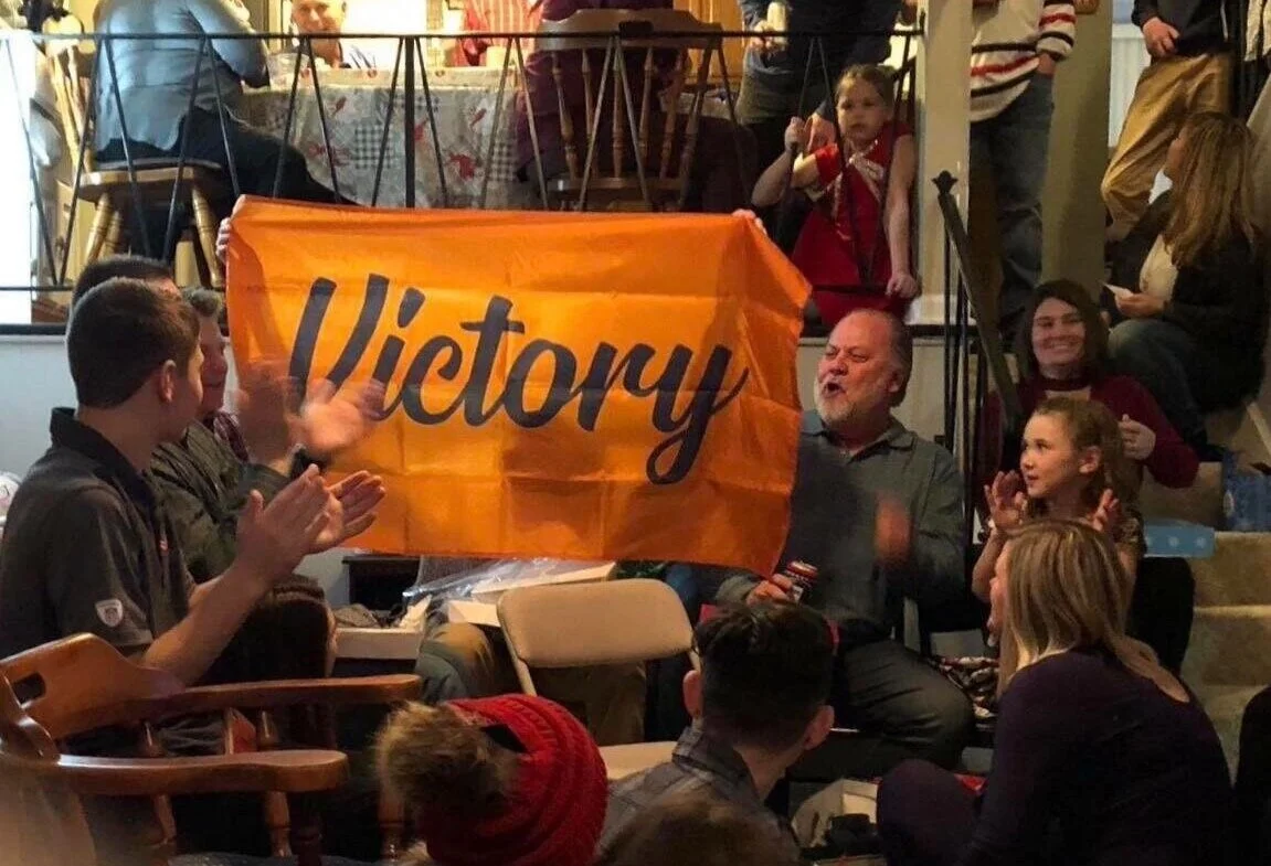 A group of people gathered indoors, with a man holding an orange flag that reads 'Victory'. Some people are clapping and smiling.