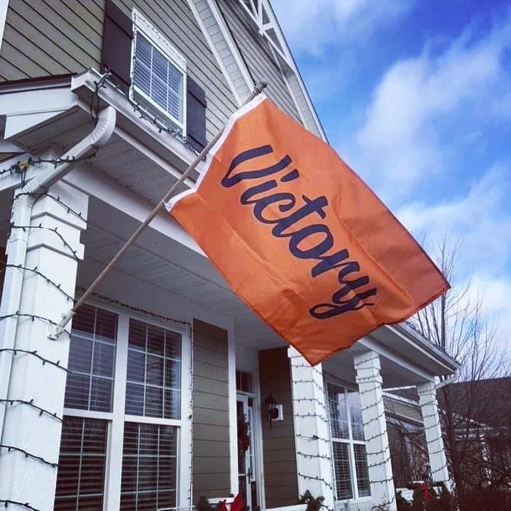 An orange flag with the word "Victory" in blue script is hanging from a pole attached to the front of a house. The house features white siding and multiple windows with blinds, under a partly cloudy sky.