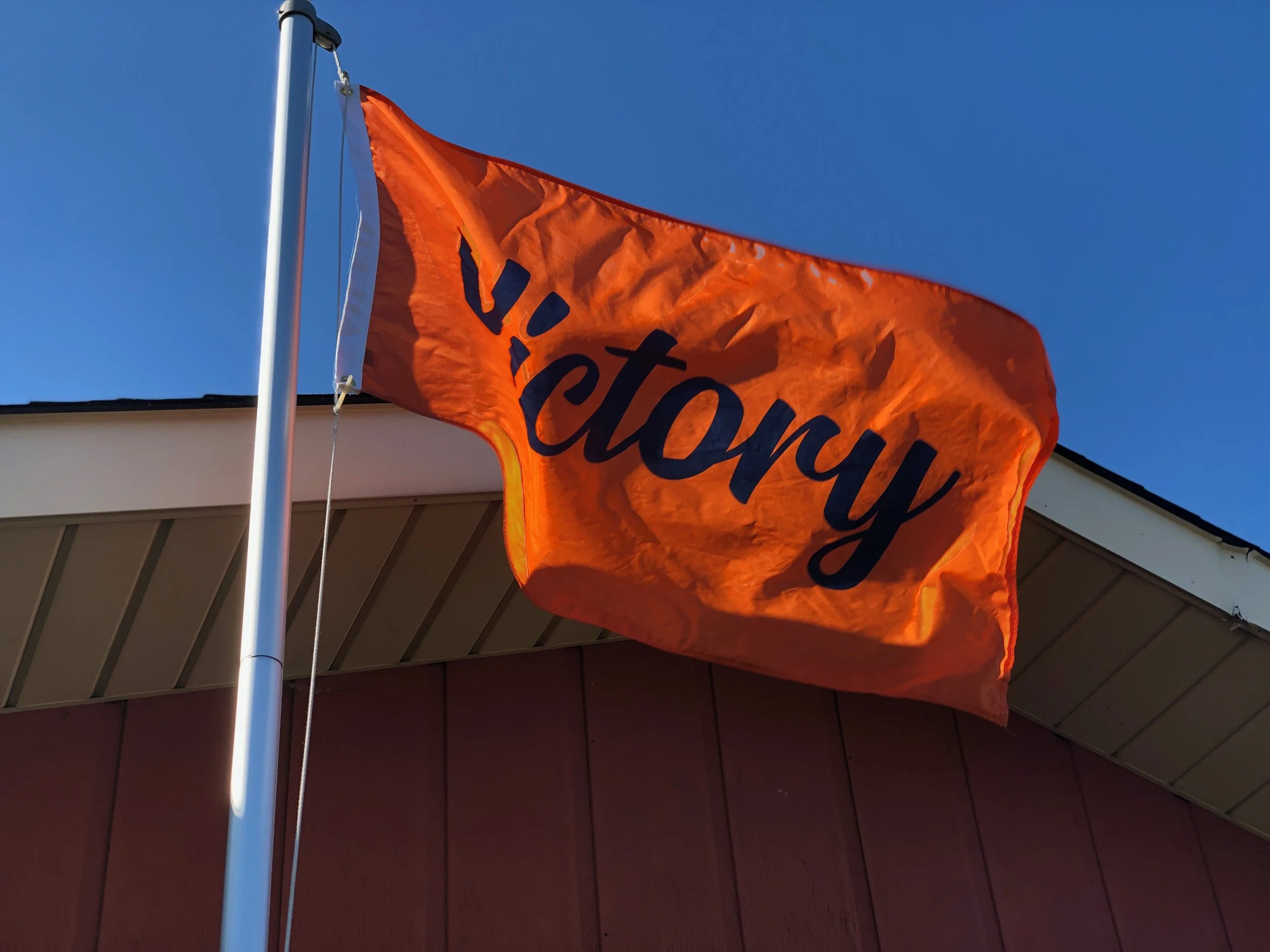 An orange flag with the word 'Victory' written in dark blue, flying against a blue sky near a building with a sloped roof.