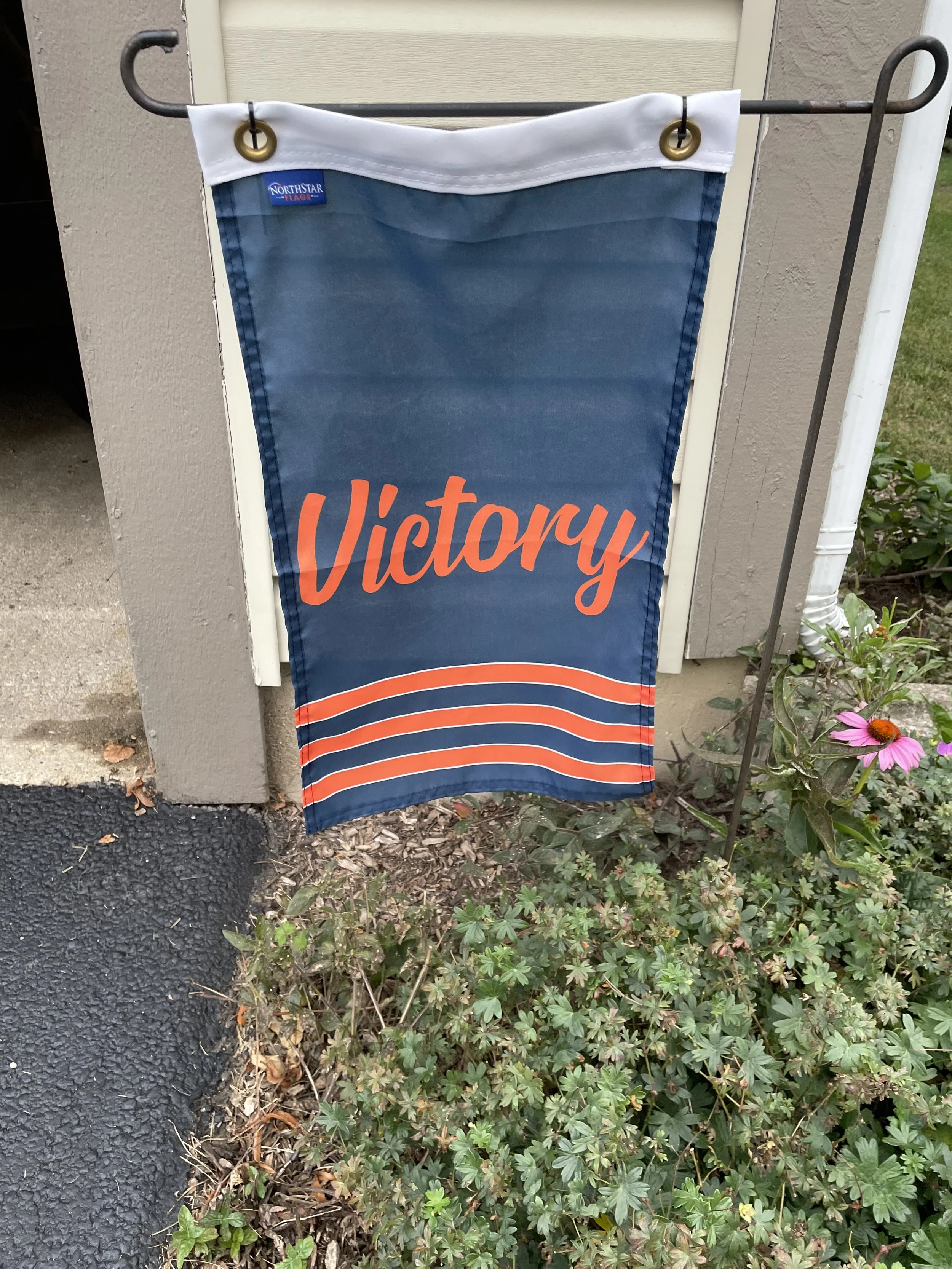 Flag with a blue background and white, red, and orange stripes hanging on a garden pole outside a house with flowers and plants.