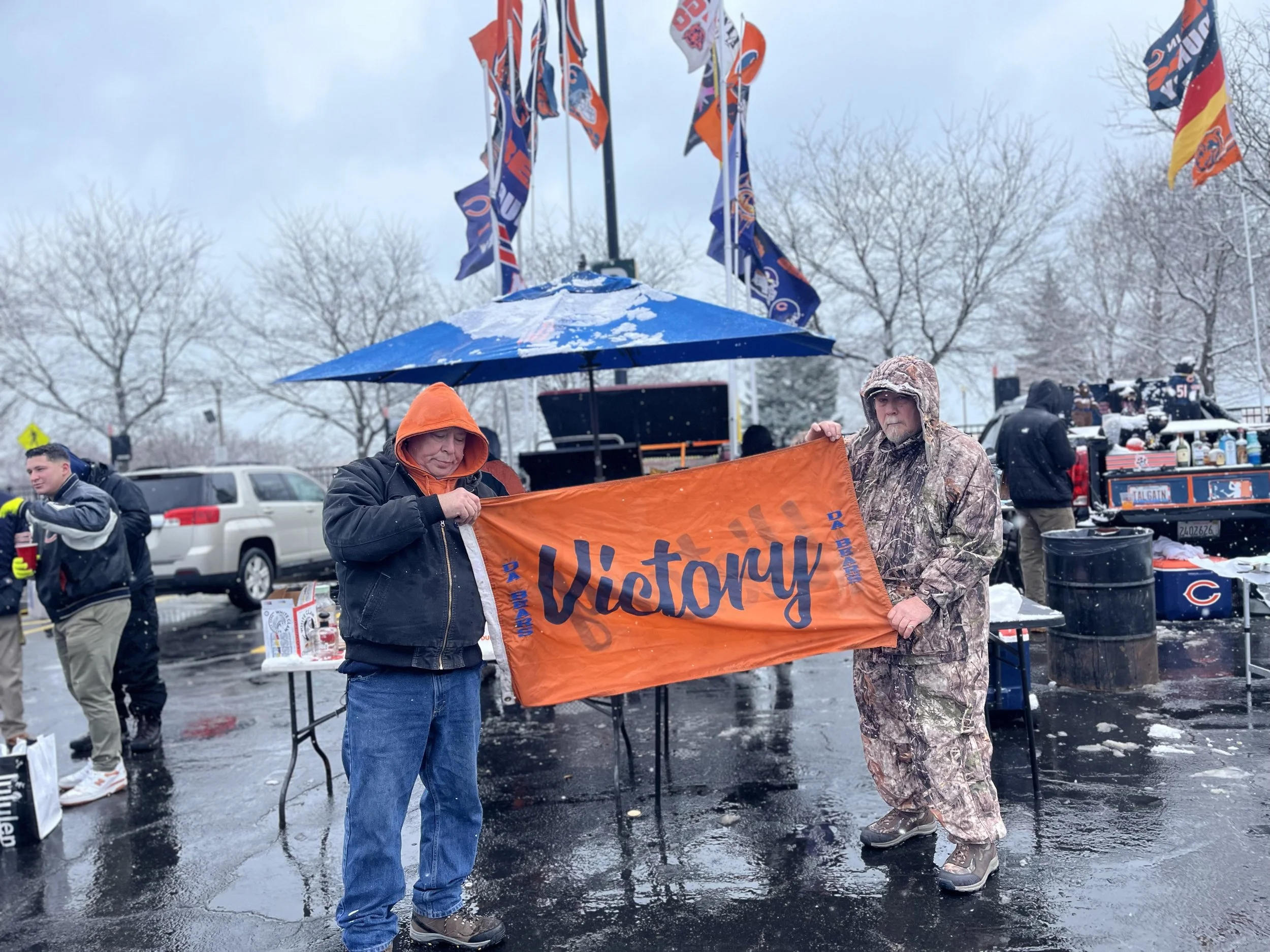 Two men are holding an orange flag with the word 'Victory' in black letters. One man is wearing a black jacket with an orange hood, and the other is dressed in camouflage rain gear. They are outdoors in a rainy parking lot with snow on the ground, an