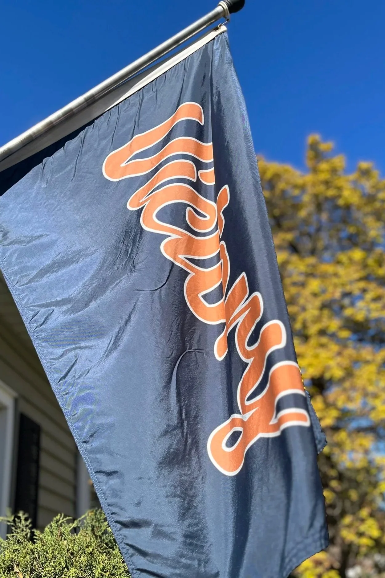 Close-up of a navy victory flag with orange and white lettering, fluttering in the wind, against a background of a tree with yellow leaves and a clear blue sky. The flag is at a chicago bears fan's house.