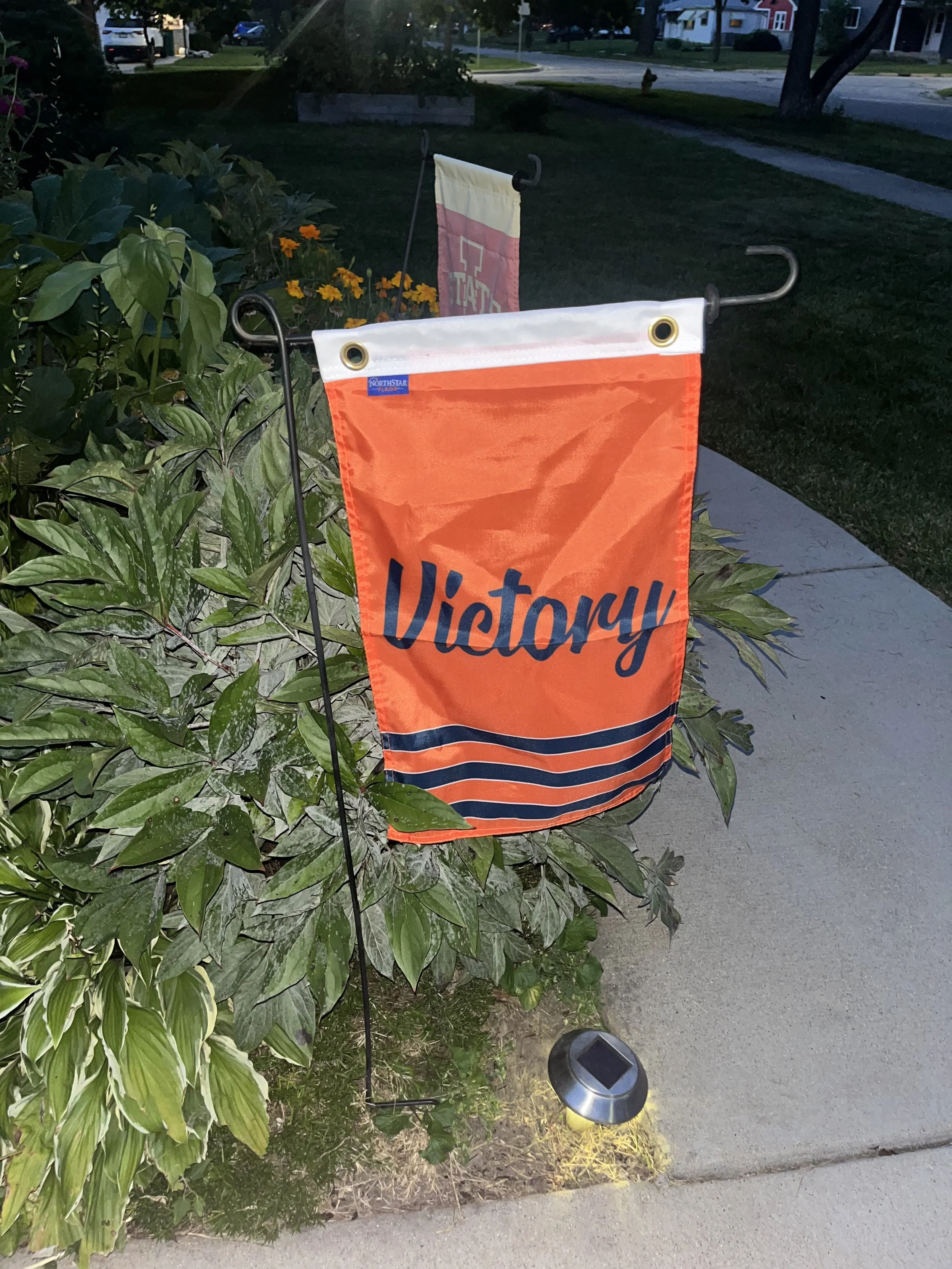 An orange garden flag on a black metal stand with the word "Victory" written on it, placed next to a green bush in a residential neighborhood at dusk, with solar lights illuminating the garden.