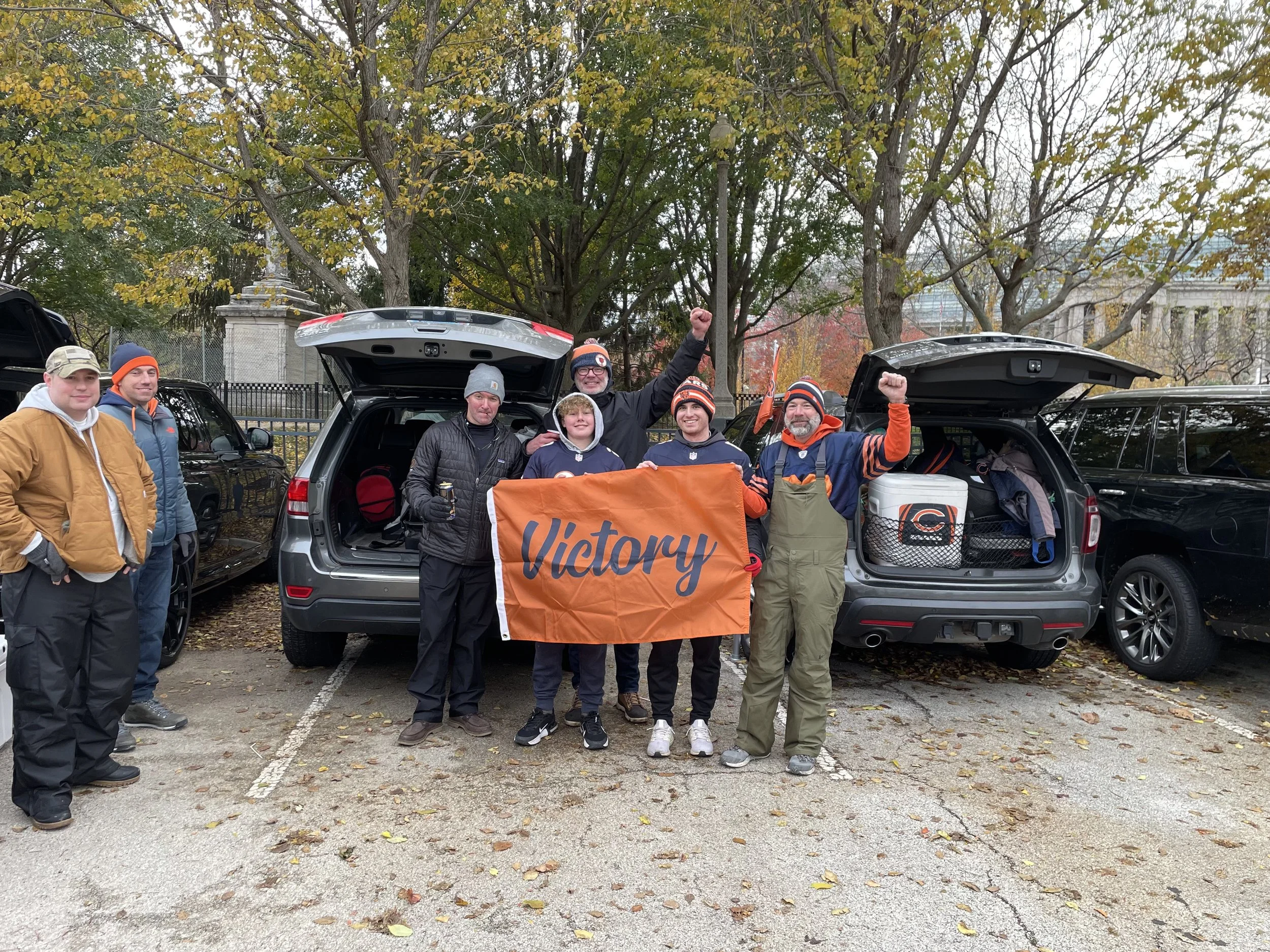 Group of men standing in a parking lot with open vehicle trunks behind them, holding a large orange flag with the word "Victory" and celebrating.