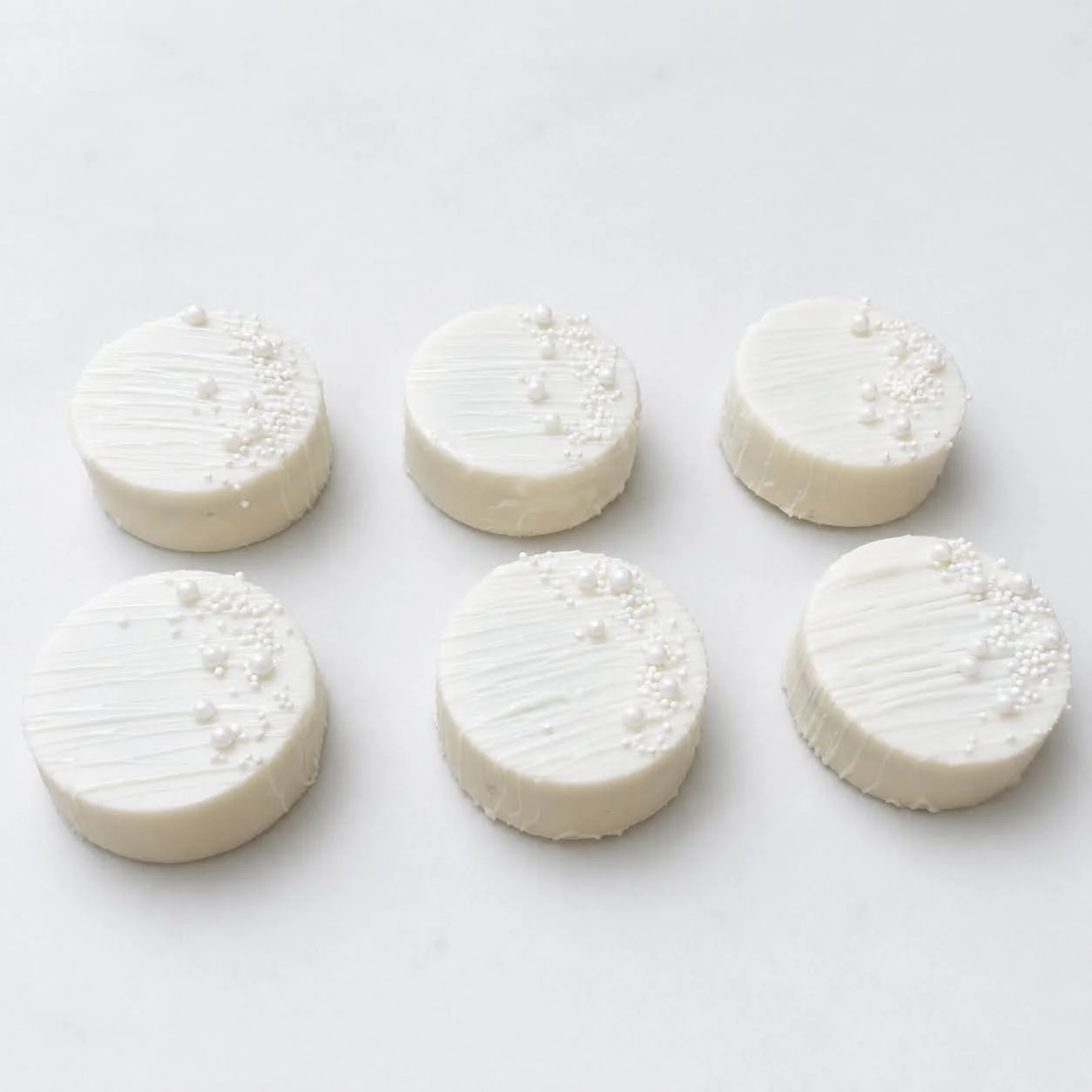 Six white chocolate-covered Oreos decorated with fine white drizzle, pearls, and small white sprinkles.