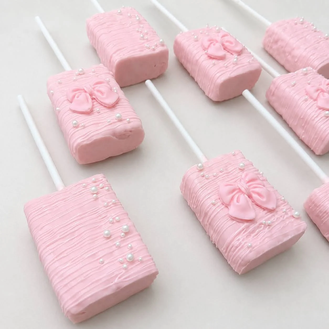 Light pink chocolate-dipped crispy treats arranged diagonally on sticks with white pearls and pink bow decorations.