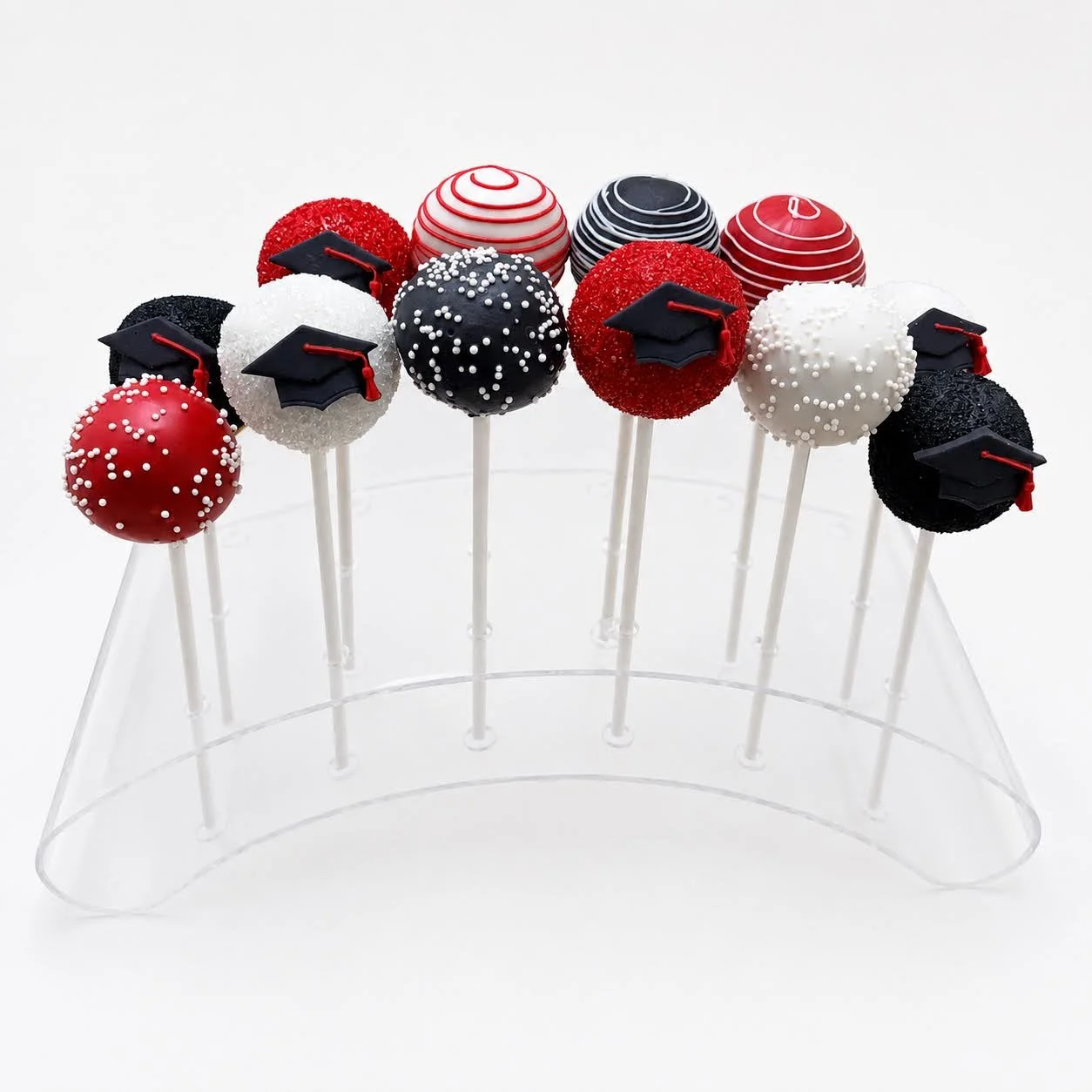 Red, black, and white graduation cake pops displayed in a clear acrylic stand with drizzle, sprinkles, and black graduation caps.