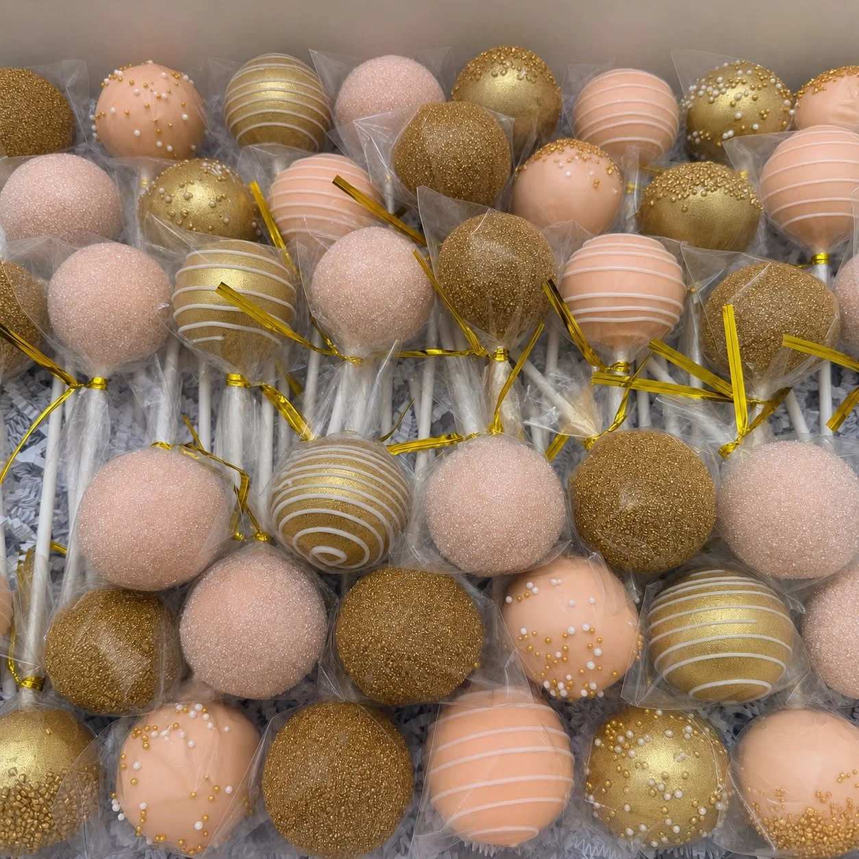 Individually wrapped peach and gold cake pops arranged in a box and finished with metallic gold ties.