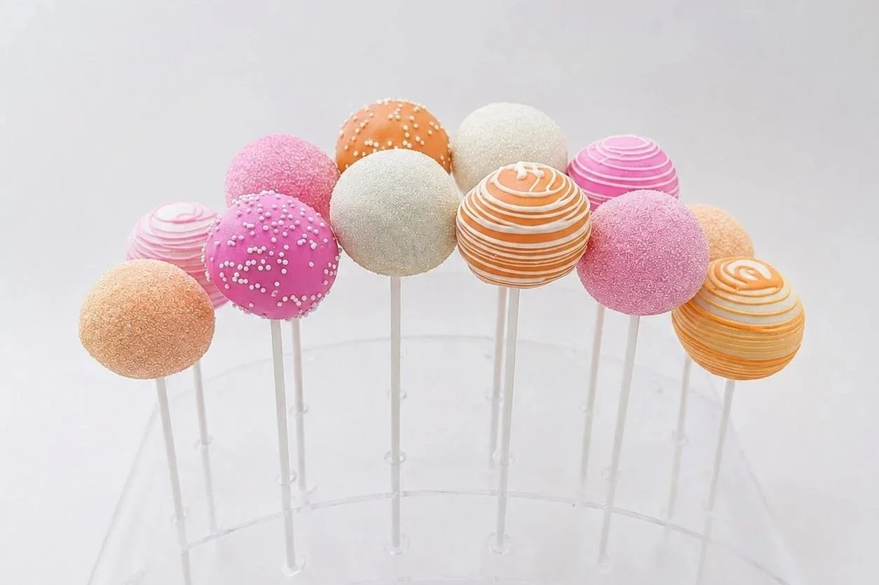 Pink, orange, and white cake pops displayed in a clear acrylic stand, decorated with drizzle, sanding sugar, and white nonpareils.