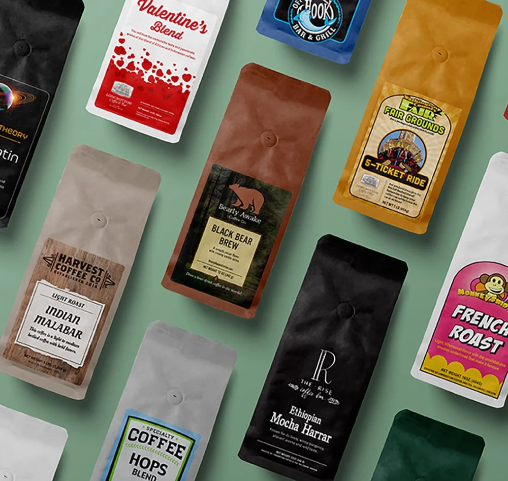 Assorted coffee bags with various labels, including Valentine’s Blend, Black Bear Brew, Indian Malabar, Ethiopian Mocha Harrar, Hops Blend, Fair Grounds 5-Ticket Ride, French Roast, and others, arranged on a light green surface.