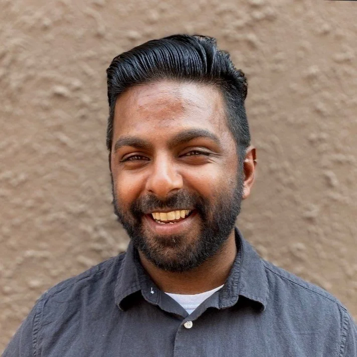 Portrait of a smiling Niladri Sinha with dark hair and beard, wearing a dark shirt, standing against a textured beige wall.