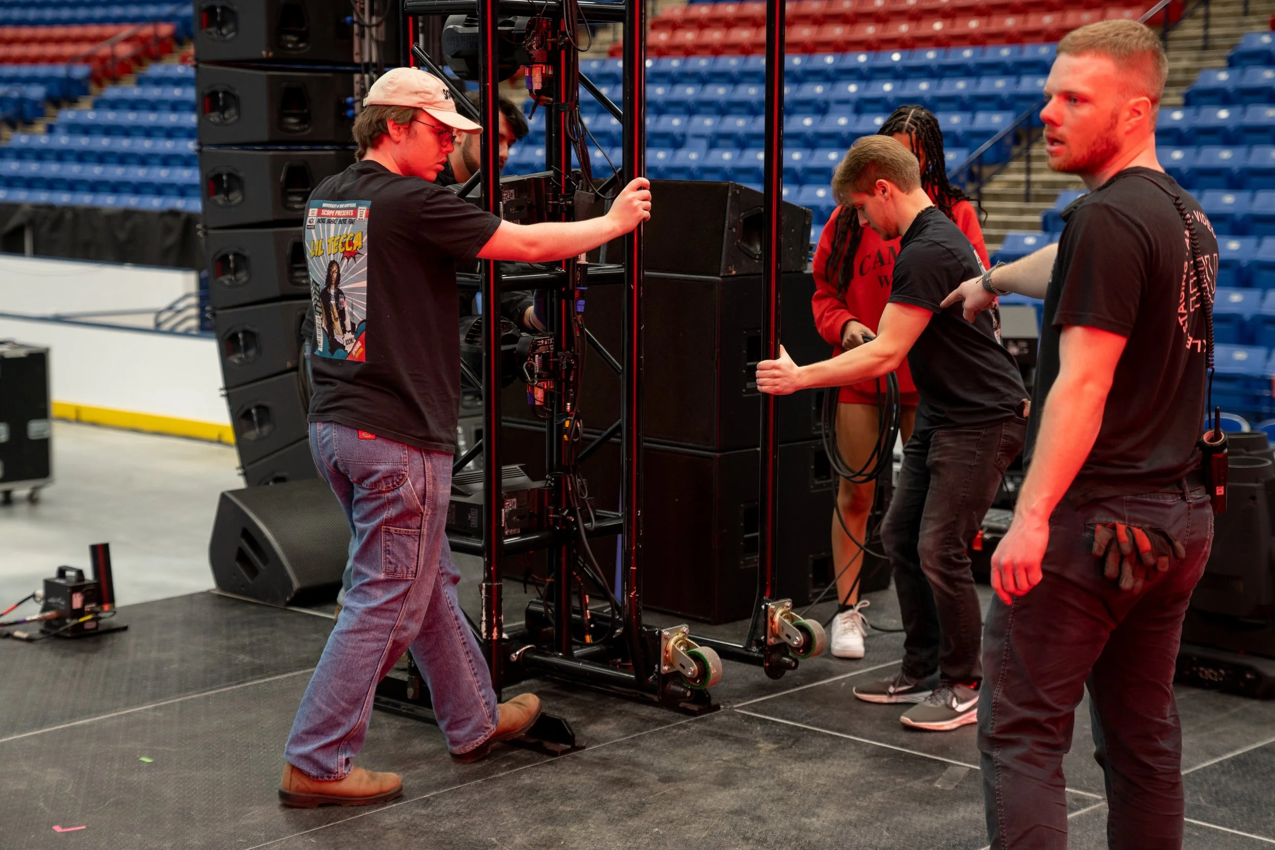 Group of five people setting up audio equipment backstage in an arena with blue seats. One man is wearing a black T-shirt with a graphic and jeans, another man with a black T-shirt, black pants, and gloves, a woman in a red hoodie, and two other men working with the black metal structure and sound equipment.