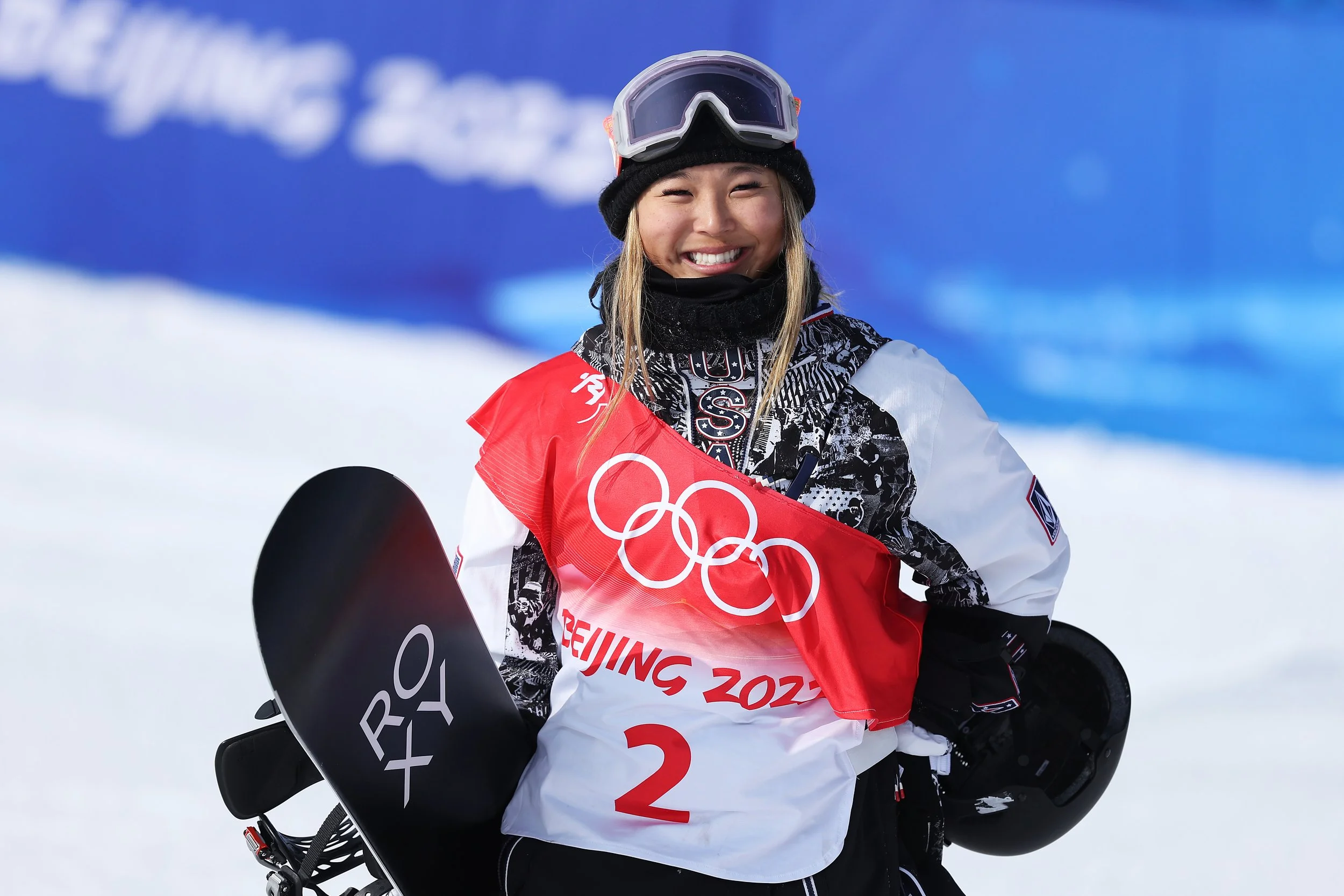 A smiling female snowboarder named Chloe Kim wearing a black beanie, goggles, and a red and white jacket with an Olympic flag and snowboard logo, holding a snowboard, at the Beijing 2022 Winter Olympics.