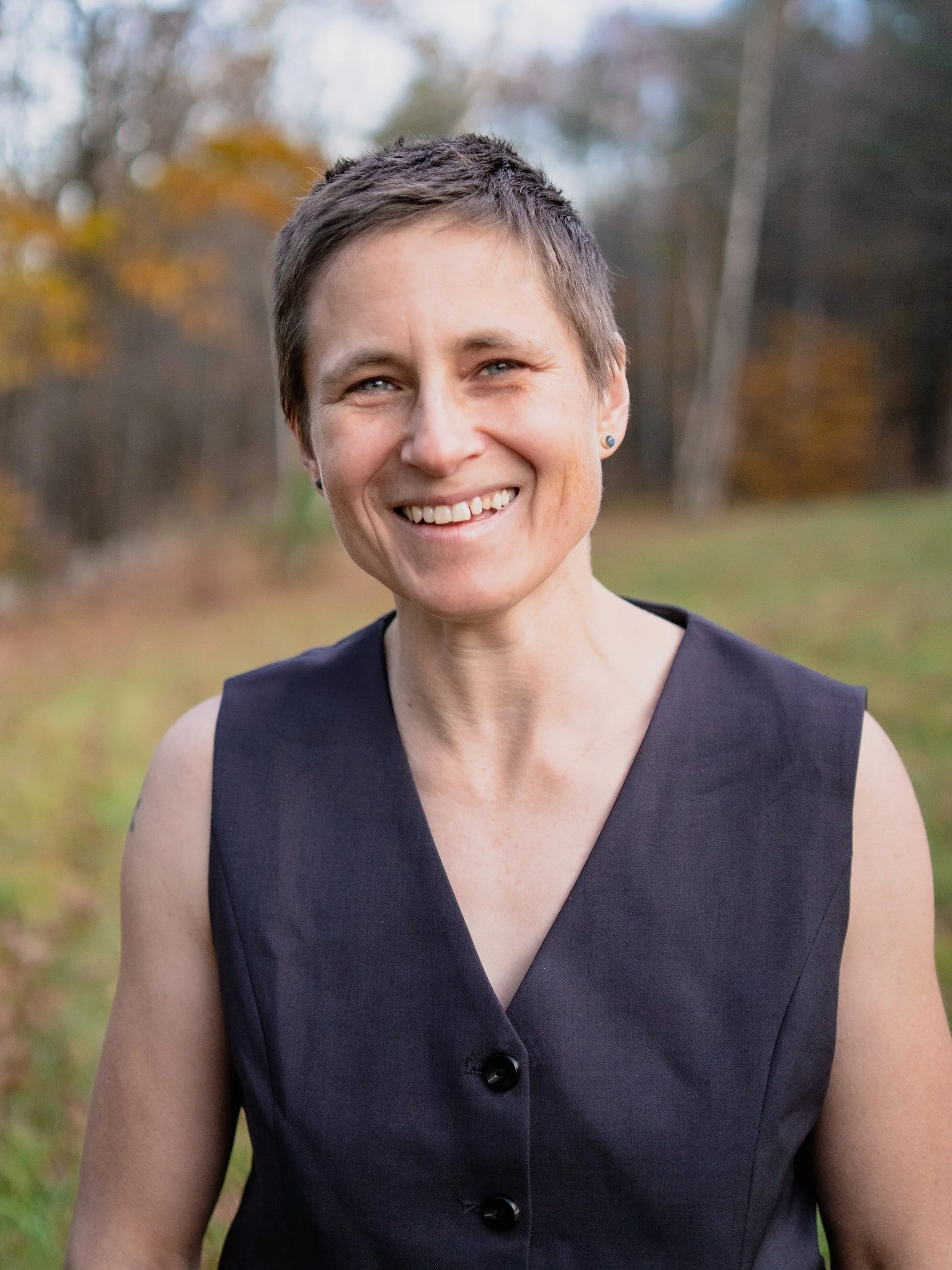 A smiling woman with short hair wearing a sleeveless black dress outdoors in a park during fall.