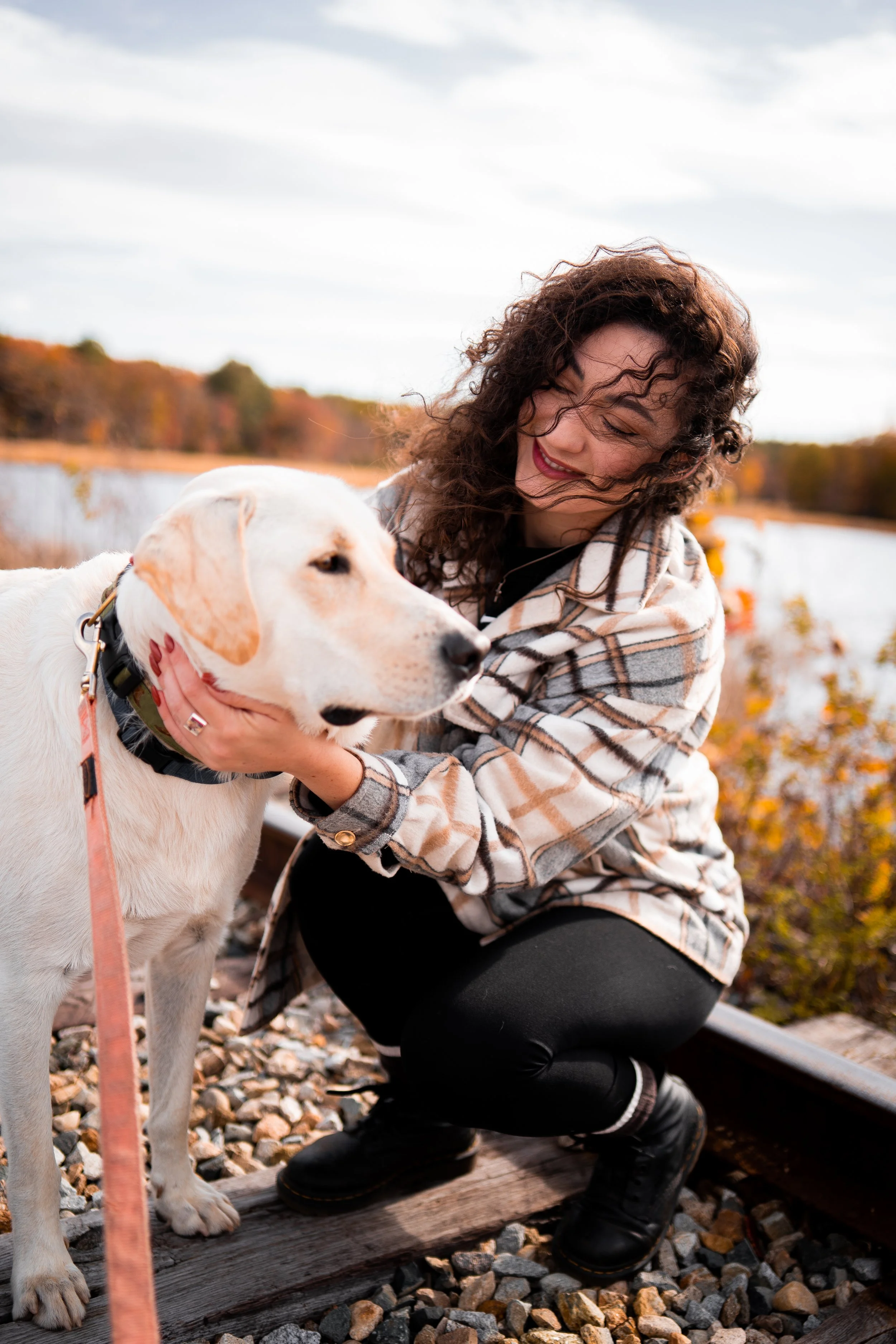 Woman with curly hair smiling while petting a yellow Labrador retriever dog beside railroad tracks in an autumn landscape.