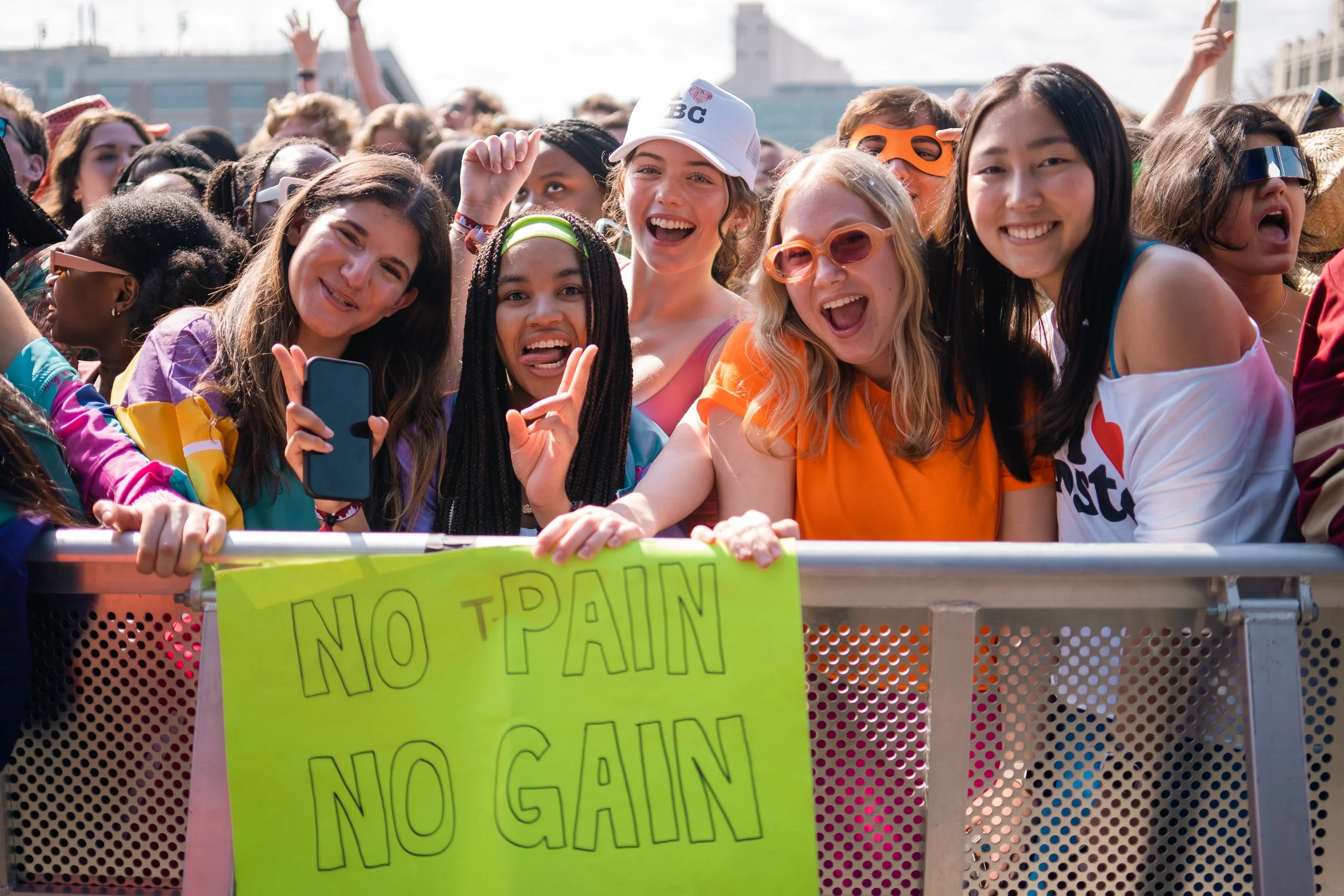 Group of diverse young women at an outdoor event, smiling and waving at the camera, holding a bright yellow sign that reads 'No Pain No Gain'.