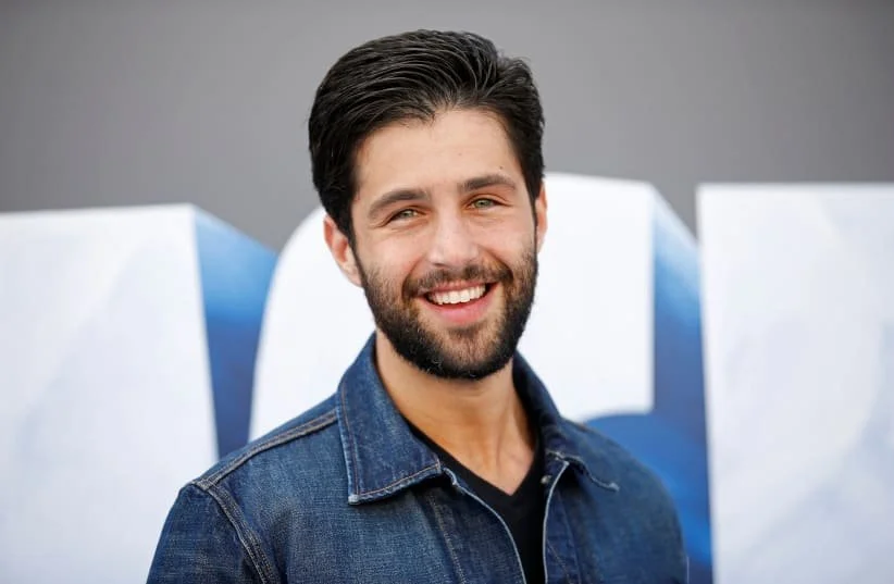 Josh Peck with dark hair and a beard, wearing a denim jacket, standing outdoors in front of white and blue sculptures.