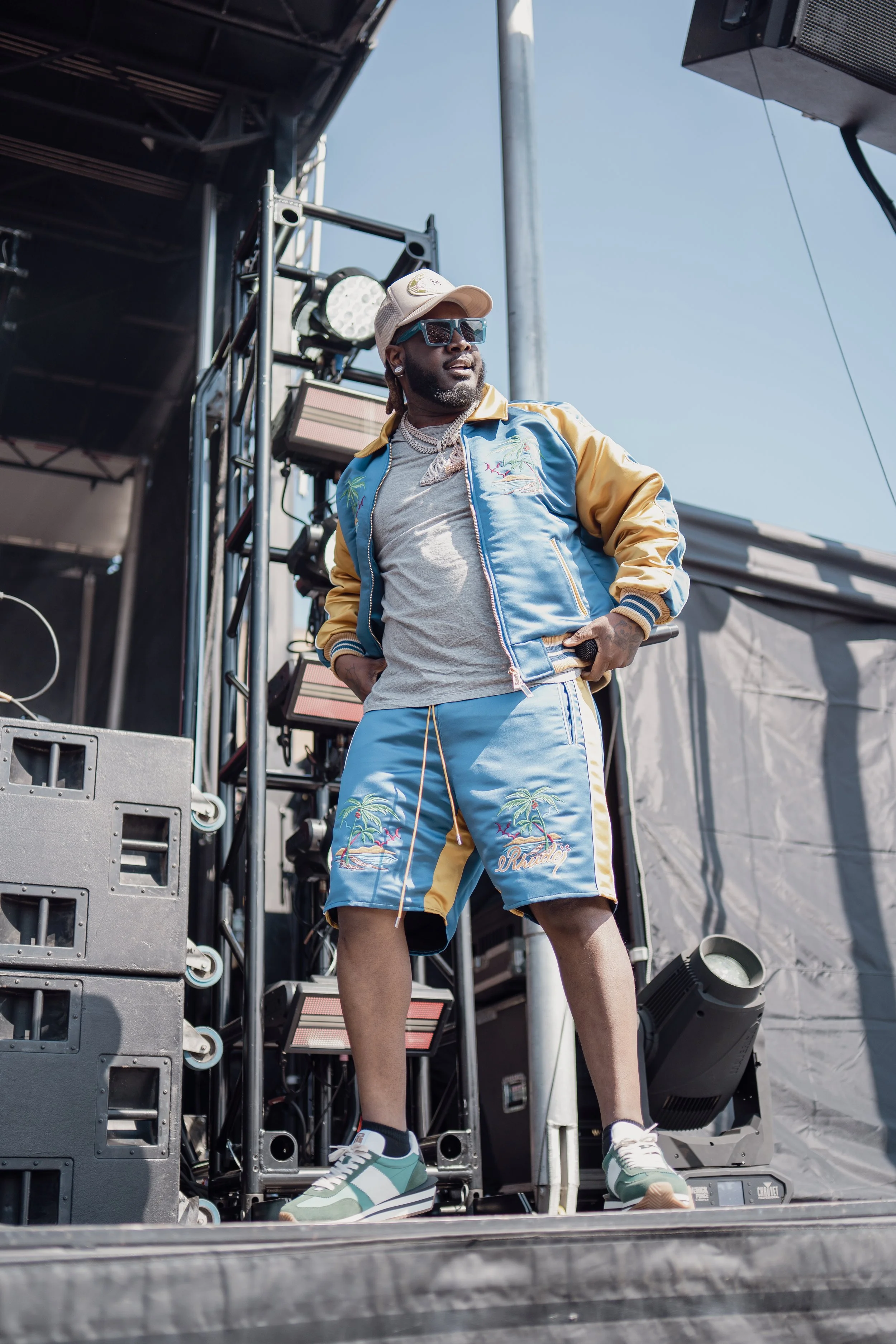 T-Pain in colorful beach-themed clothing, including a blue and yellow jacket and shorts with palm tree and island embroidery, standing on a stage with sound equipment and lighting, under a clear blue sky.