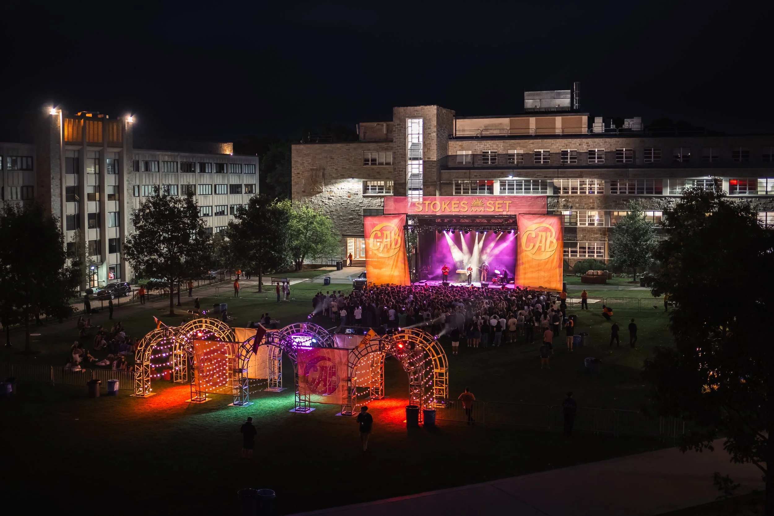 An outdoor night concert at Boston College with a stage on a lawn, colorful lights, and a crowd of people watching. Surrounding trees and buildings are visible in the background.