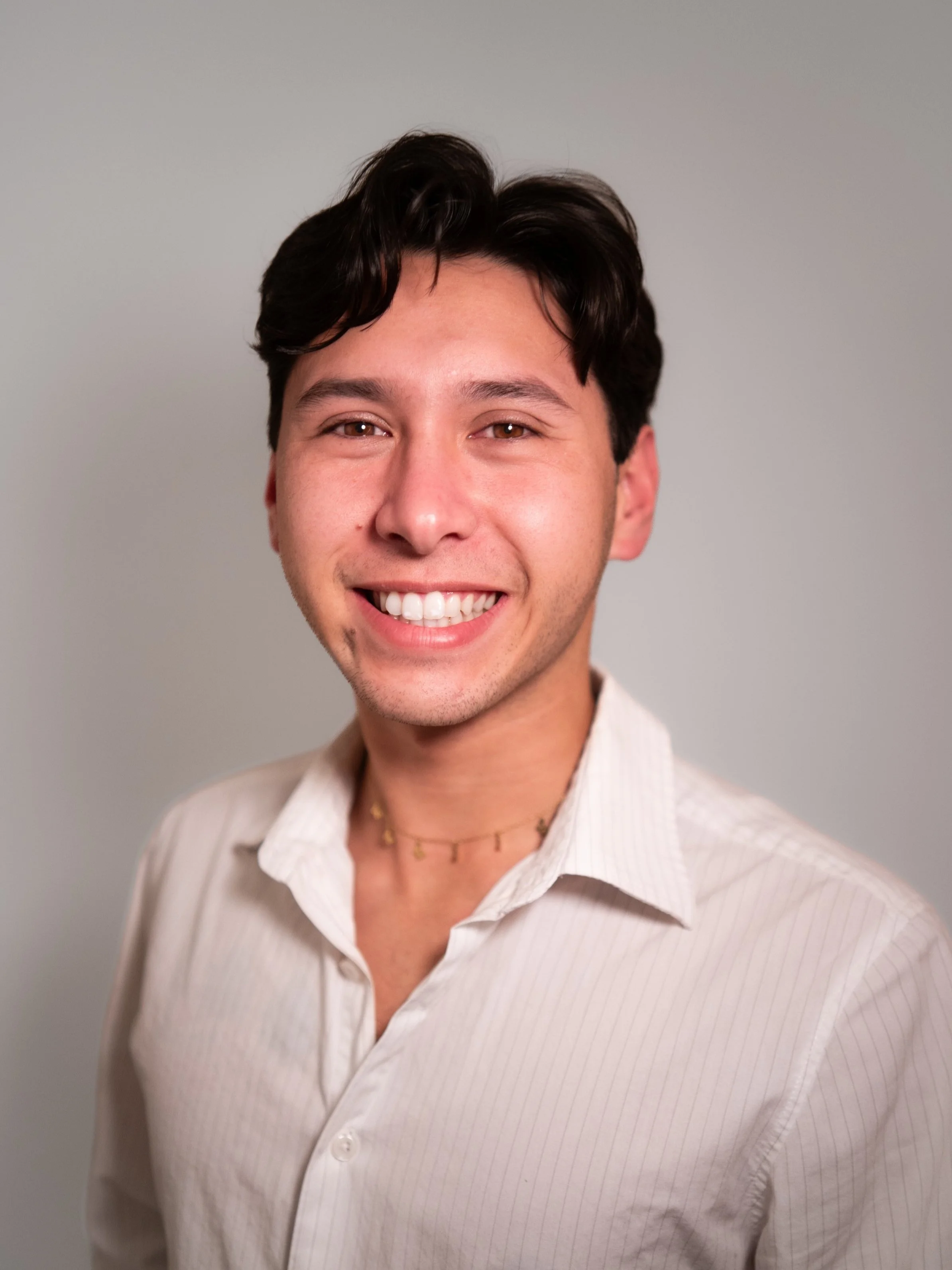 A young man with dark hair and a light complexion, smiling in a white dress shirt, standing against a plain background.