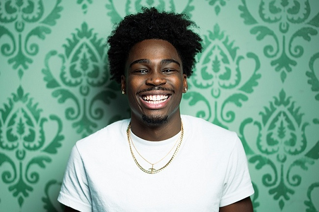 Kam Patterson wearing a white shirt, gold chains, and a small earring, standing in front of a green decorative patterned wallpaper.