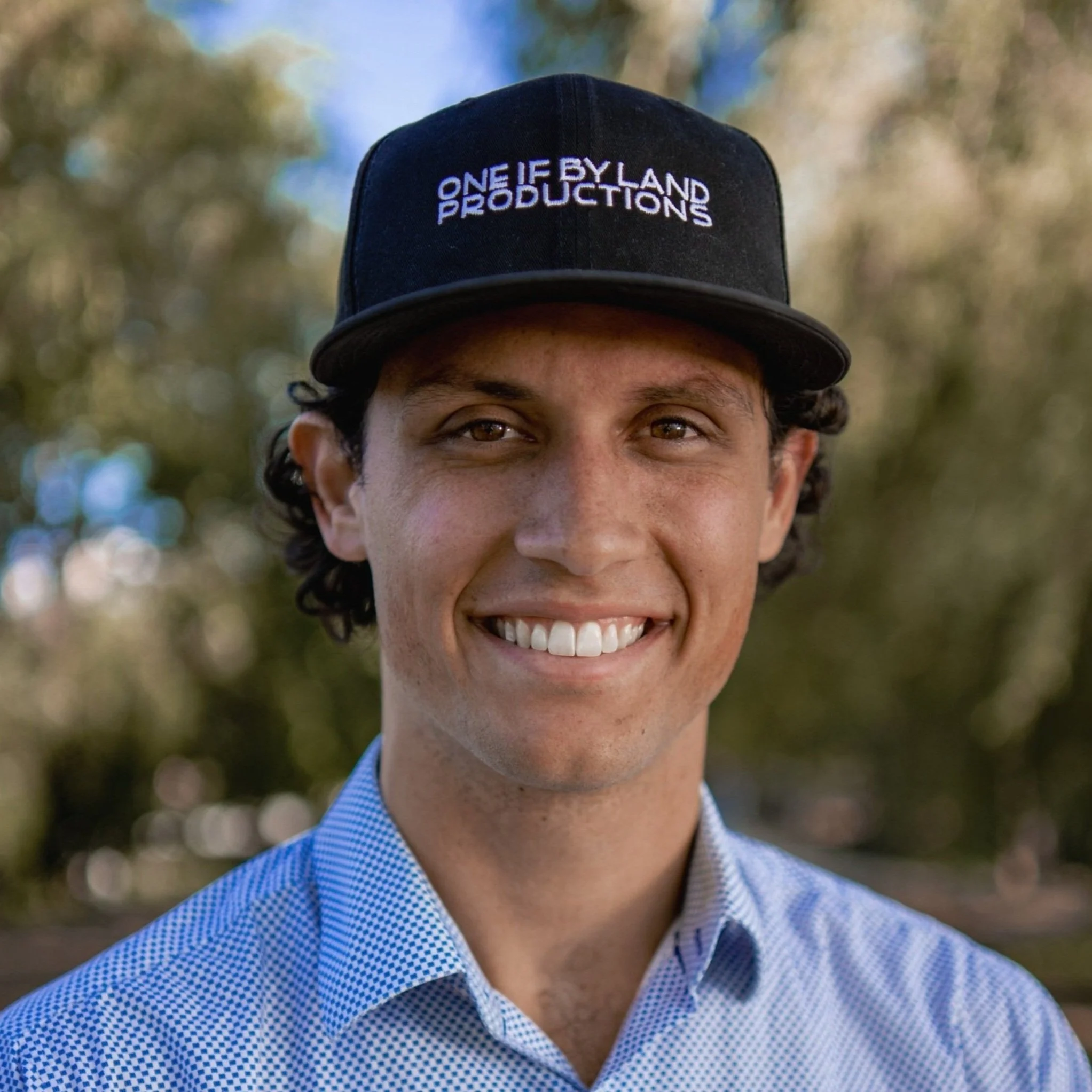 A smiling Harrison Flagg with curly dark hair wearing a black baseball cap with the text 'One If By Land Productions' and a light blue checkered shirt, standing outdoors with trees in the background.