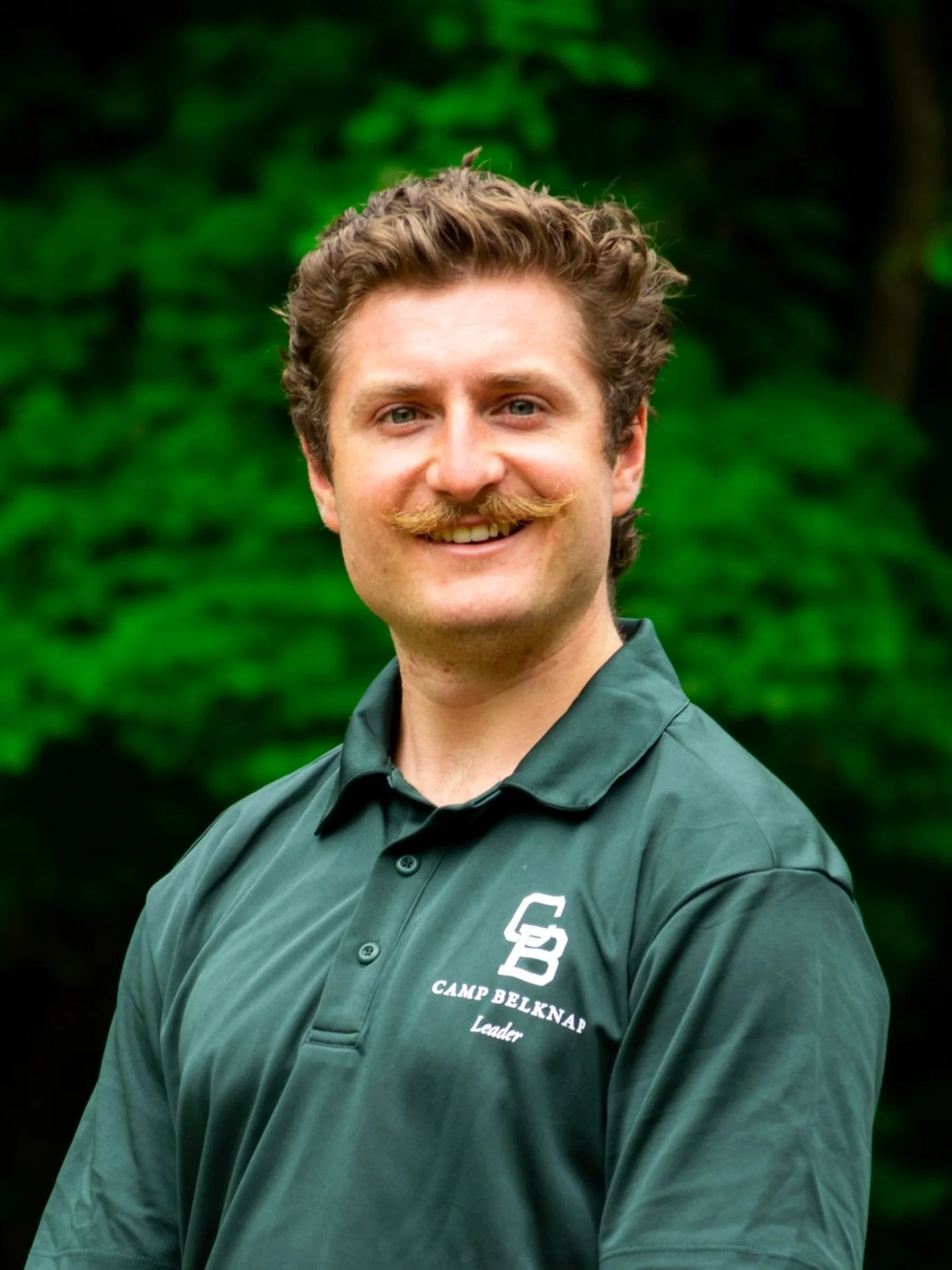 A man with curly hair and a mustache wearing a green polo shirt with 'Camp Belknap' and 'Leader' embroidered on it, standing outdoors in front of green trees.
