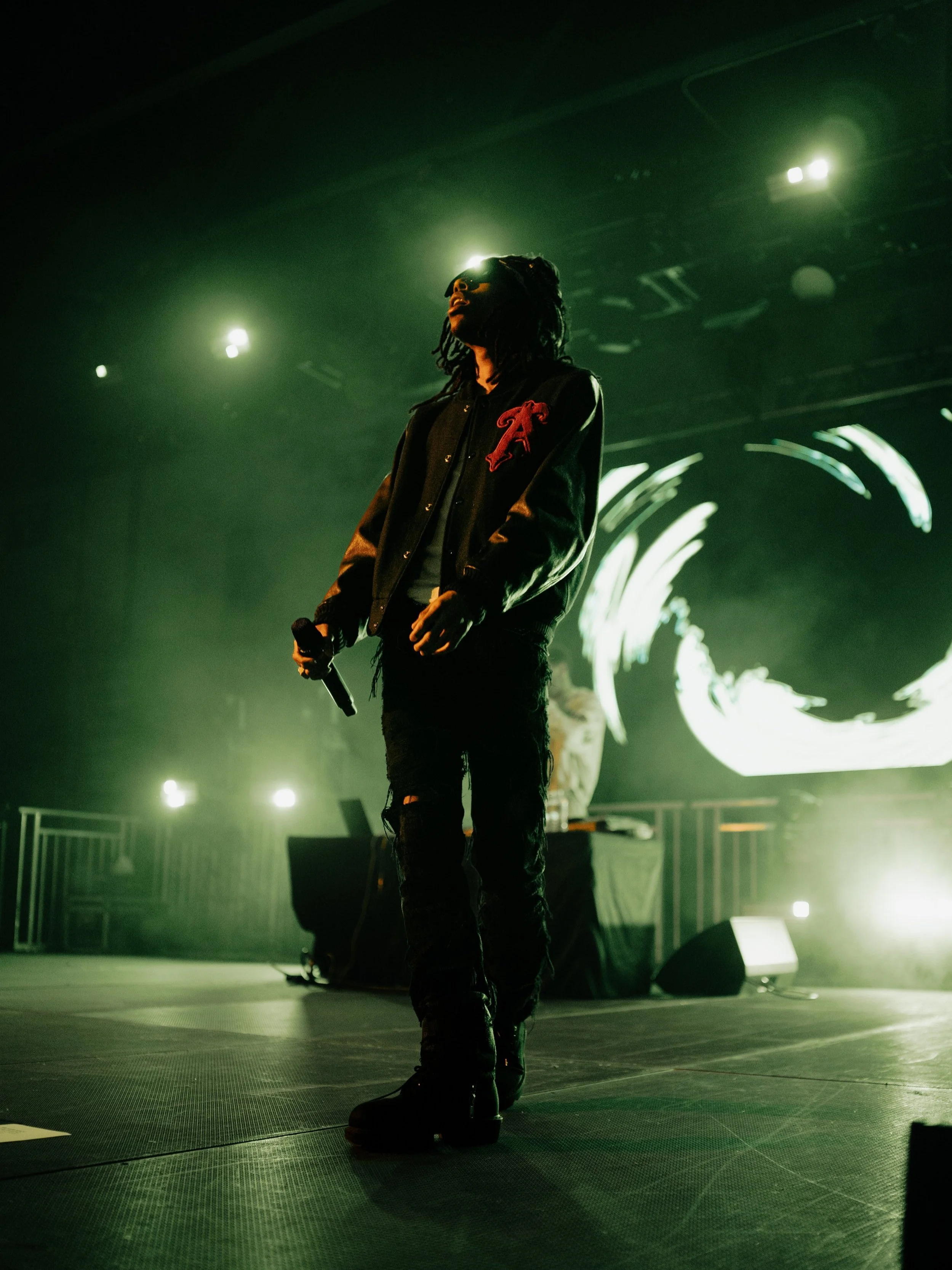 A performer stands on stage with a microphone in hand, illuminated by stage lights, in front of a large digital screen displaying abstract graphics.